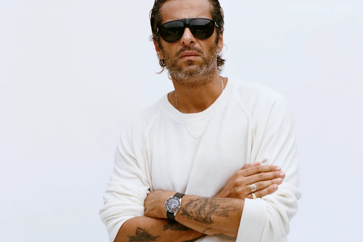 A man with his arms crossed wearing dark District Vision Yukari Windshield wraparound sunglasses in a six base frame.
