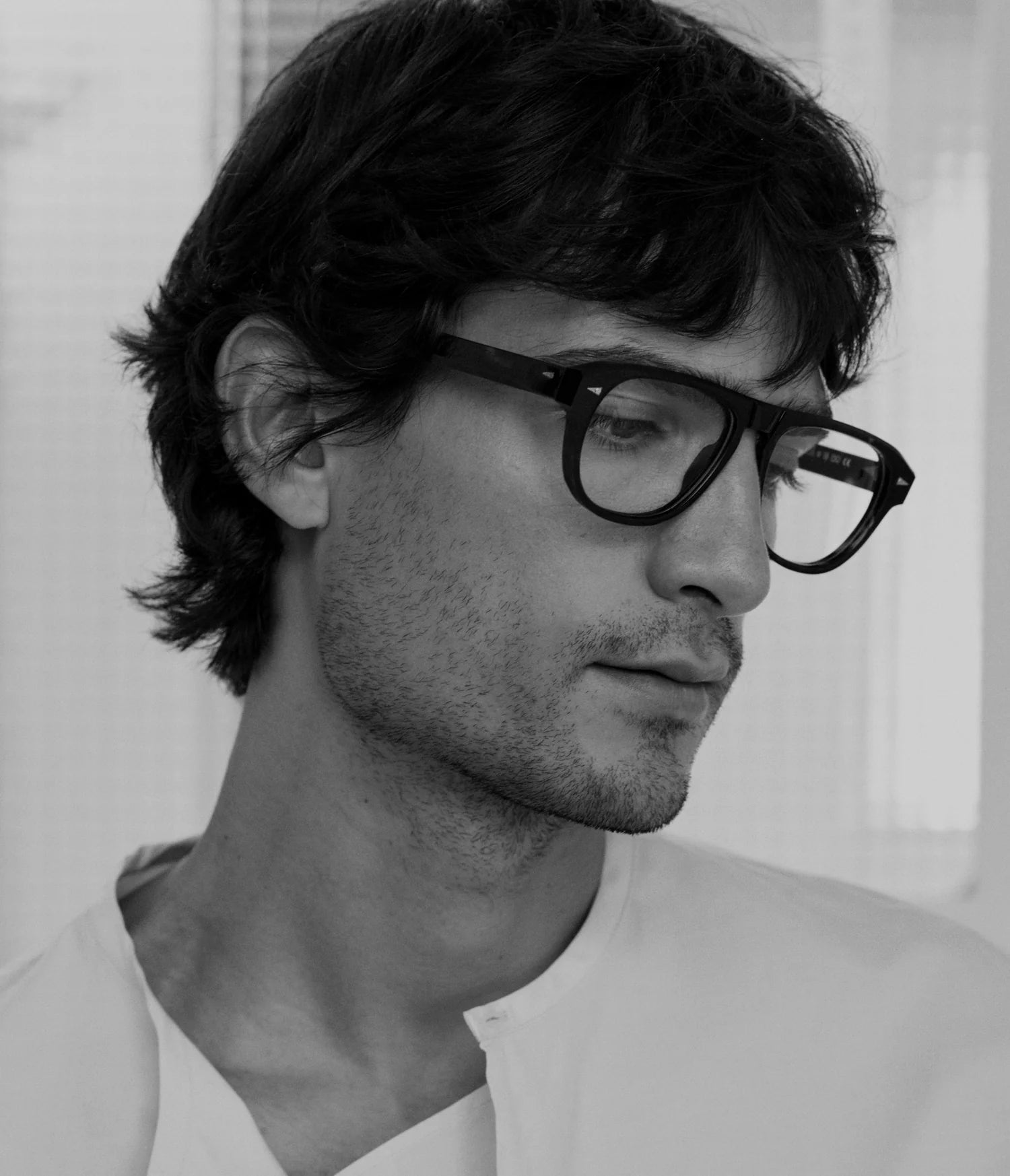 A black and white portrait of a man with dark hair and a light stubble, wearing thick black eyeglasses and a white shirt.