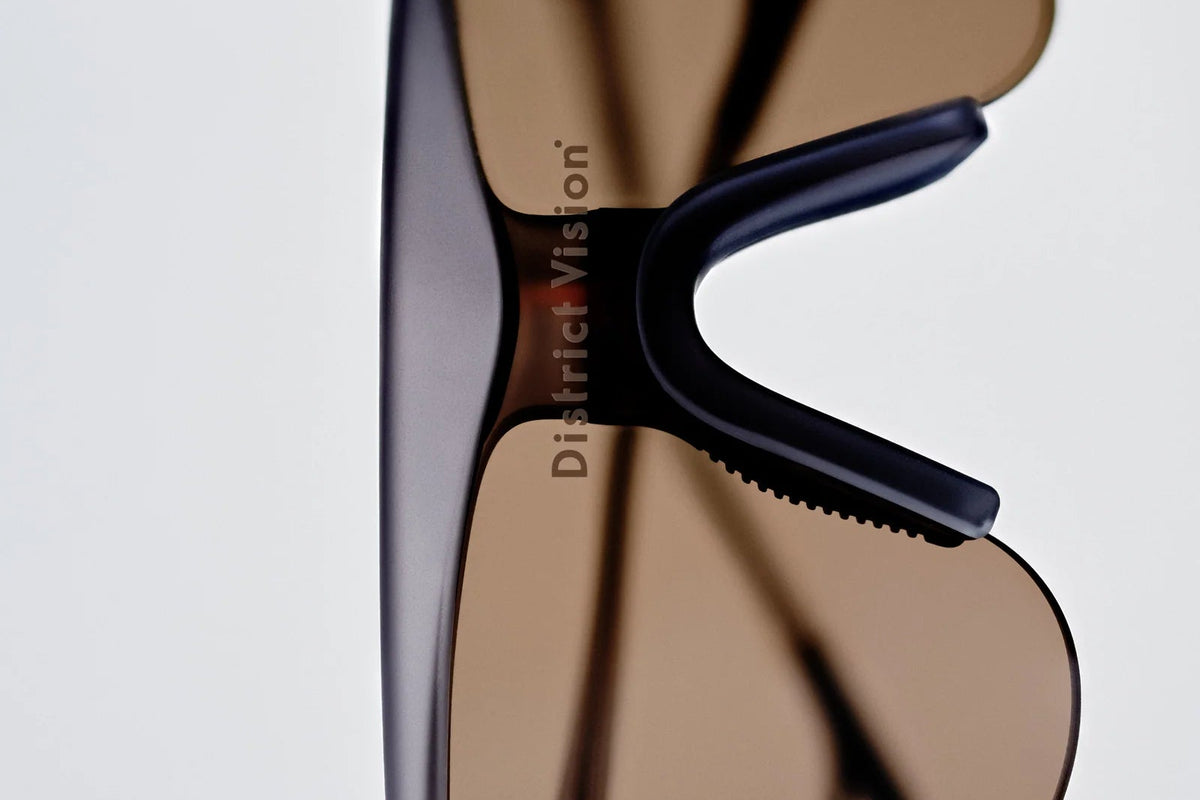 Close-up of the District Vision Momoka Pacer sunglasses, featuring brown-tinted, therapeutic grade lenses and a translucent gray TR90 frame. The District Vision logo appears on the lens, while the softly lit plain background highlights the ergonomic design.