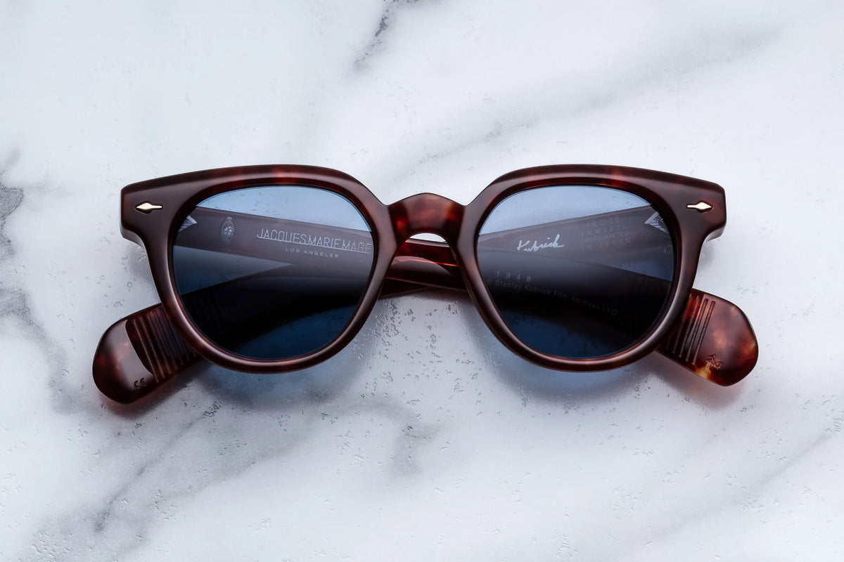 Tortoiseshell-patterned acetate sunglasses with blue-tinted lenses from the Stanley Kubrick by JMM: 1948 collection resting on a white marble surface.