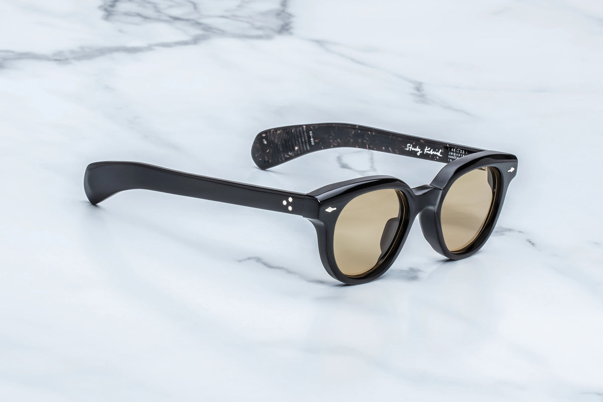 Black thick-rimmed Stanley Kubrick by JMM: 1948 sunglasses with amber lenses and patterned inner temples displayed on a white marble surface as a cinematic tribute to iconic limited-edition eyewear.