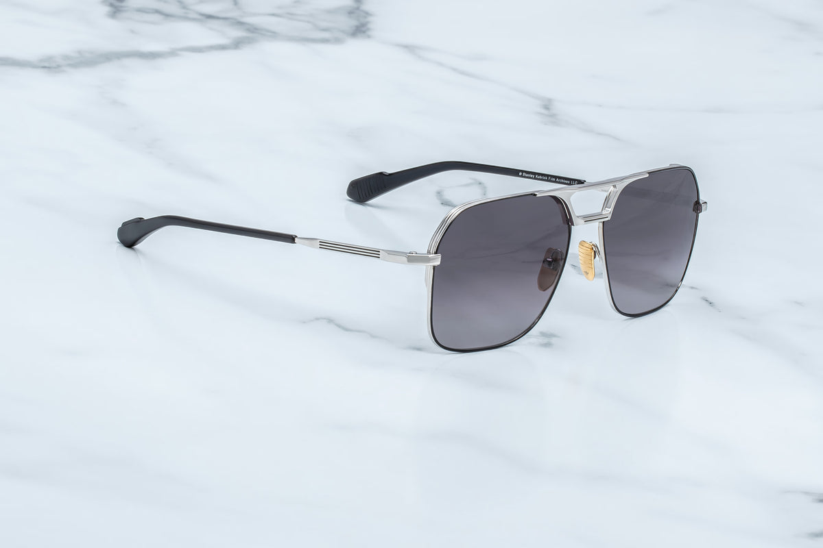 The Jacques Marie Mage Stanley Kubrick by JMM: 1980 sunglasses—aviator-style shades in silver and black with dark lenses, inspired by the iconic eyewear of Stanley Kubrick—rest on a white marble surface.