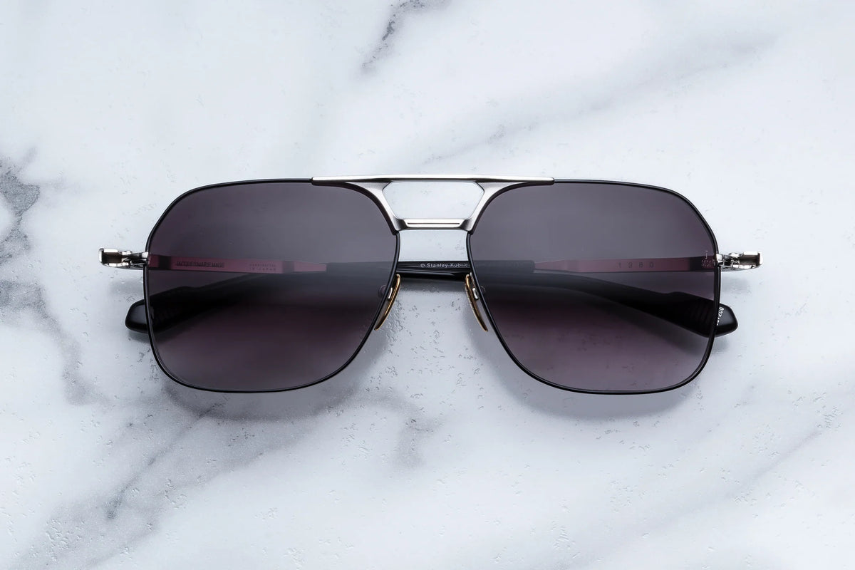 A pair of Jacques Marie Mage “Stanley Kubrick by JMM: 1980” black aviator sunglasses with dark lenses and metal frames rests on a white marble surface, evoking a subtle filmmaker tribute inspired by Stanley Kubrick’s iconic eyewear.