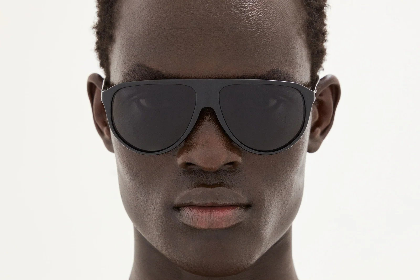 A person with dark skin and short curly hair wears the District Vision Yukari Windshield sunglasses in a bold aviator style from District Vision, looking directly at the camera against a plain, light background.