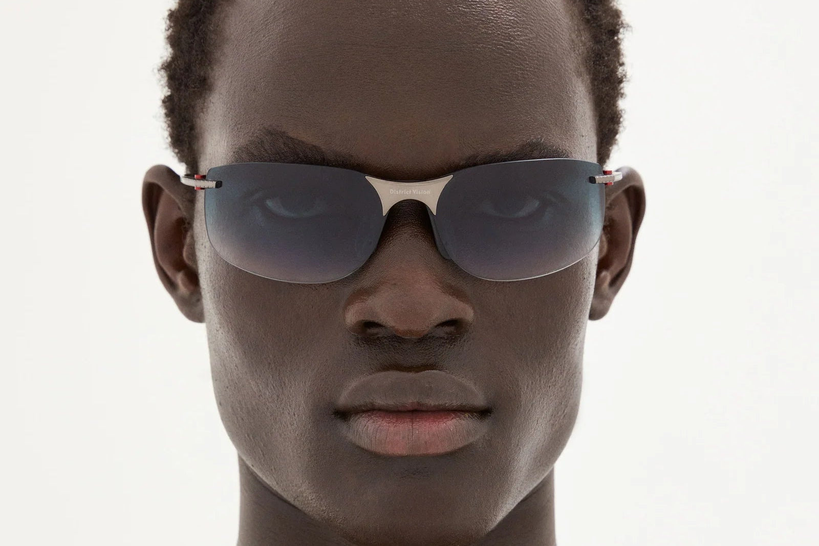 Wearing District Vision Miho Windstream Ti sunglasses by District Vision, a person with short curly hair looks directly at the camera against a plain light background.