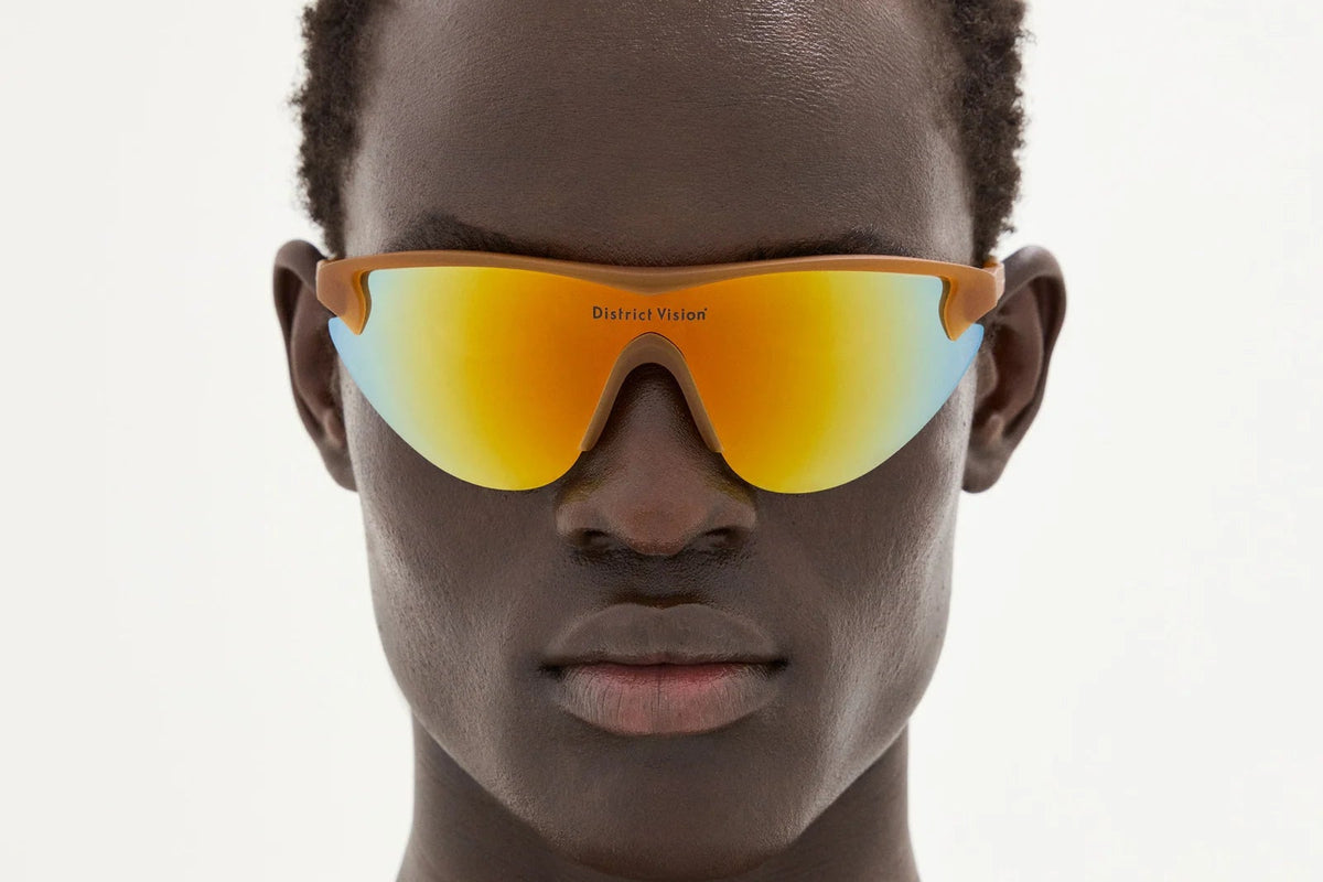 A person with short hair wears the District Vision Yukinori Range Finder, orange mirrored running sunglasses from District Vision—renowned for their high-performance cycling eyewear—against a plain light background.