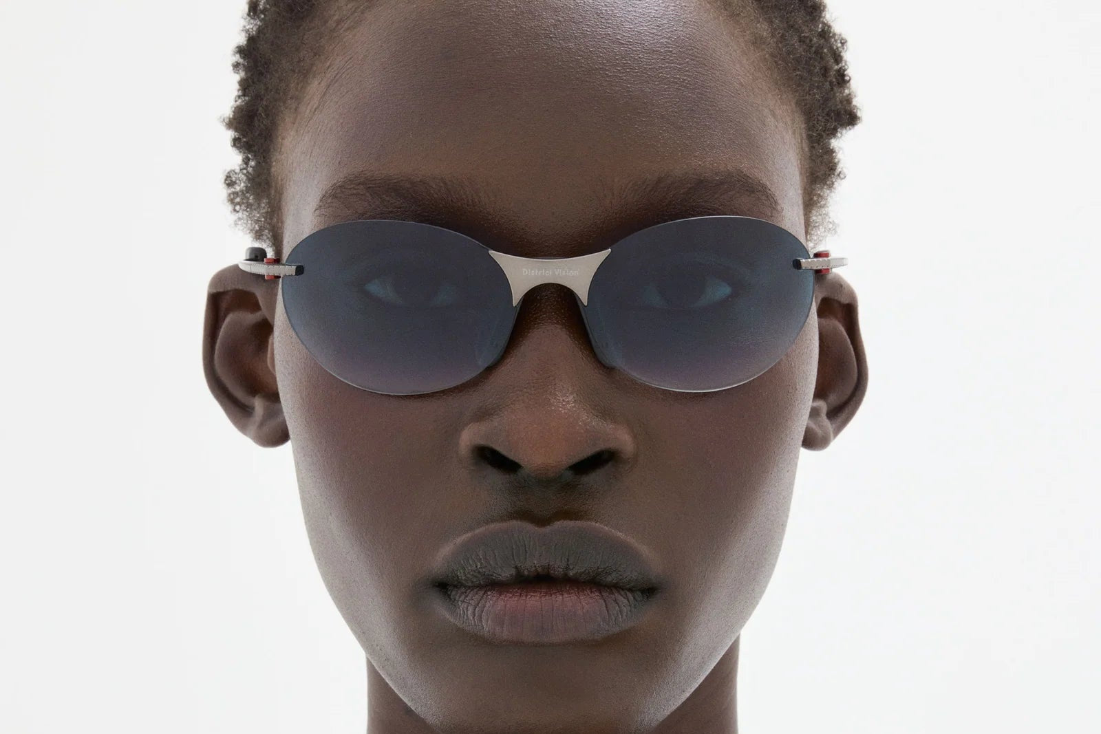 A person with short curly hair wears District Vision Mami Vantage Ti sunglasses by District Vision, featuring titanium temples, along with a black tank top, looking straight at the camera against a plain light background.
