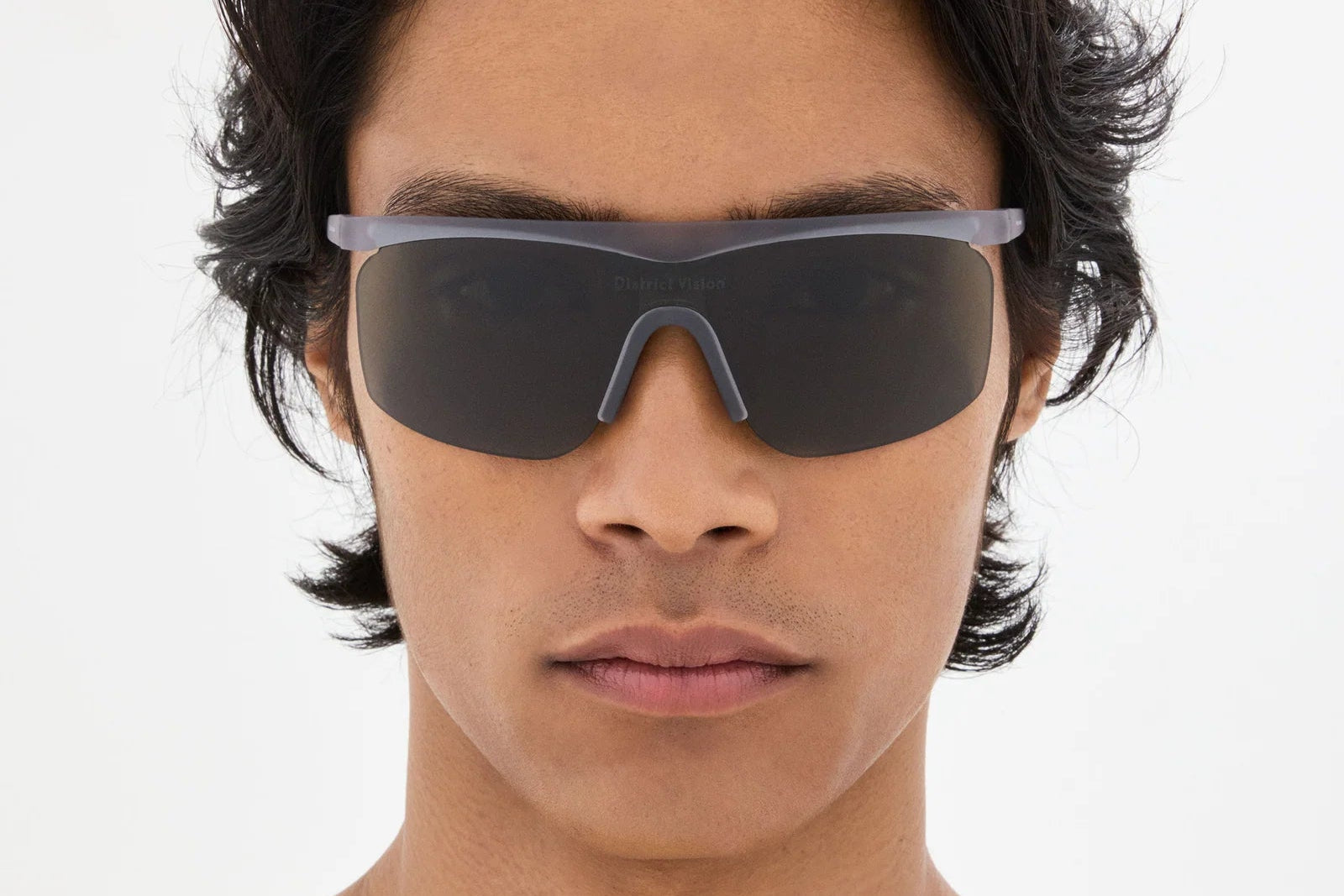 A person with wavy dark hair wears the District Vision Konoha Sun Sprinter sunglasses by District Vision, featuring large, futuristic black rectangular blade lens frames enhanced by D+ lens technology, and looks directly at the camera against a plain, light background.