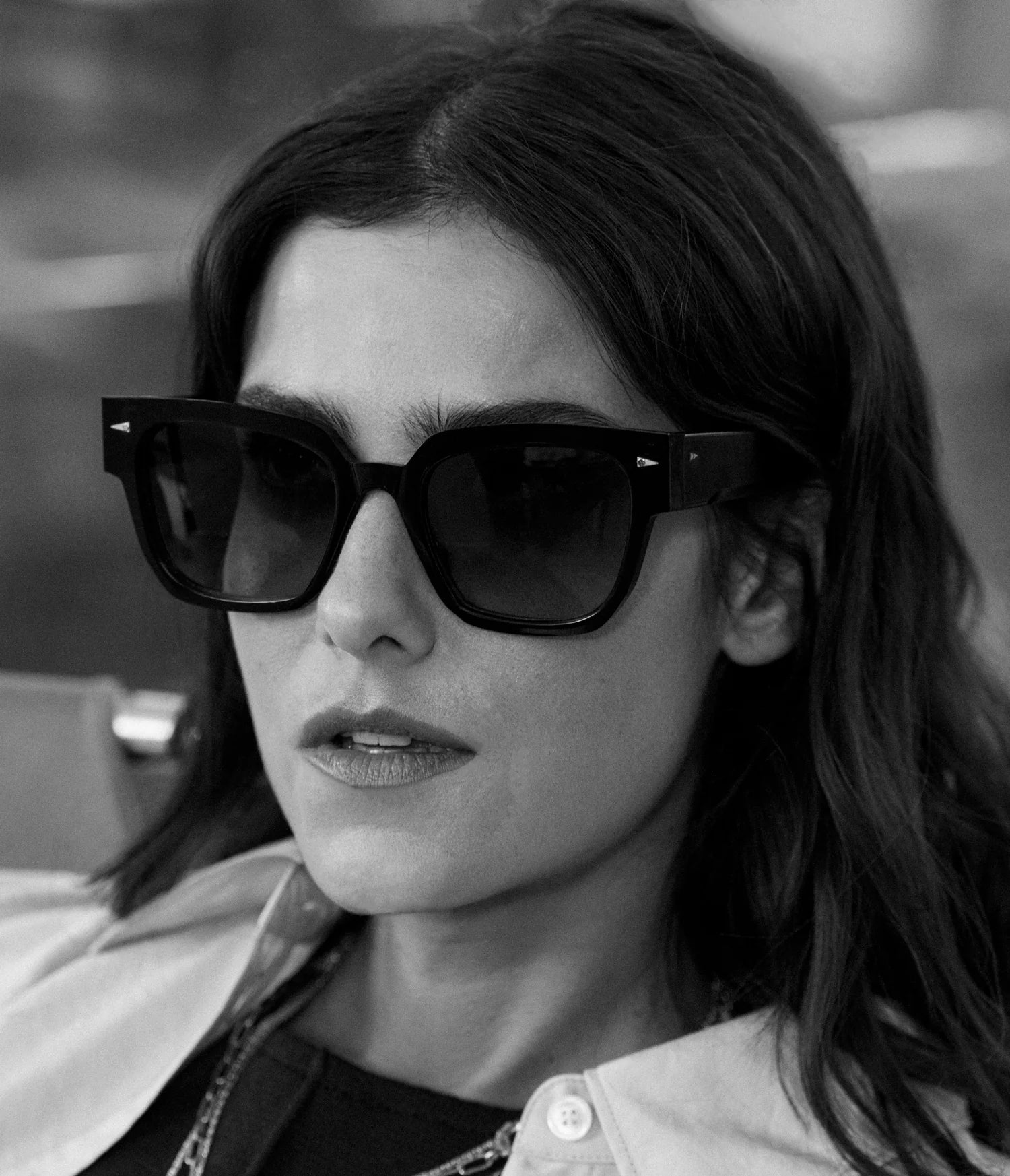 A woman with wavy dark hair wears oversized AHLEM Italiens handcrafted French sunglasses and a light-colored shirt over a dark top. She looks serious and gazes slightly to the side. The photo is black and white.