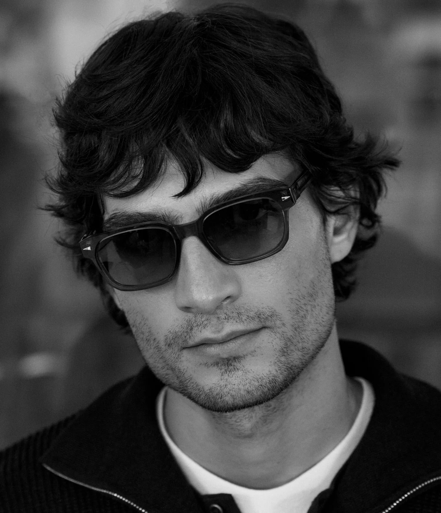 A man with wavy dark hair, light stubble, and dressed in a dark jacket over a light shirt gazes slightly away from the camera. He is wearing AHLEM Sedaine luxury French eyewear by AHLEM in this black-and-white photograph.