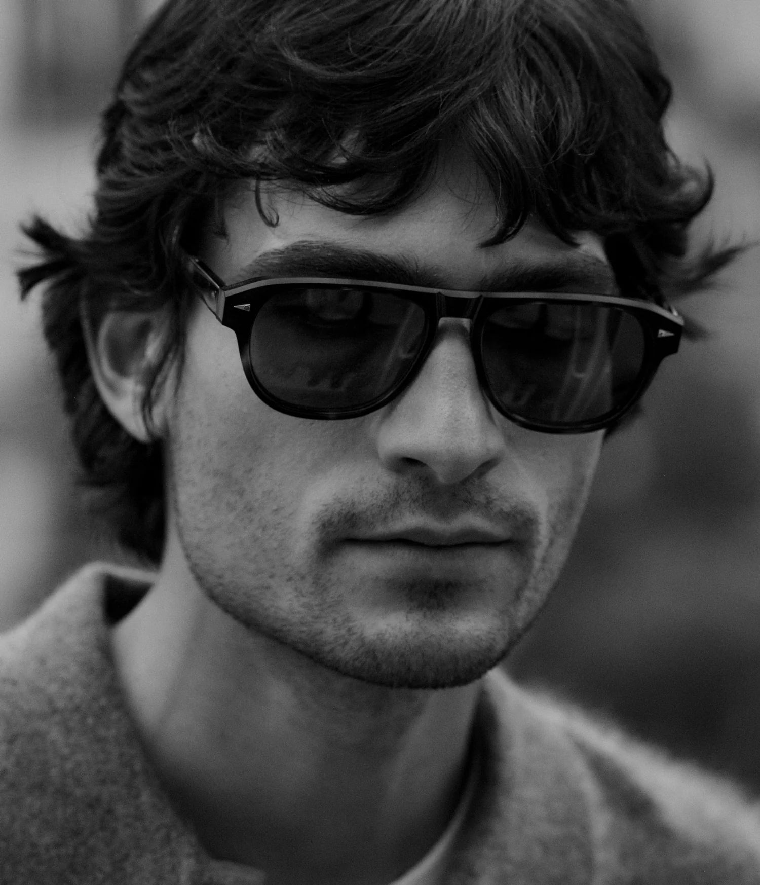 A man with wavy hair wearing AHLEM Carpentier pilot-shaped sunglasses and a coat is shown in a black and white close-up portrait. The premium Italian acetate frames from AHLEM, handcrafted in France, add a touch of sophistication to his look.