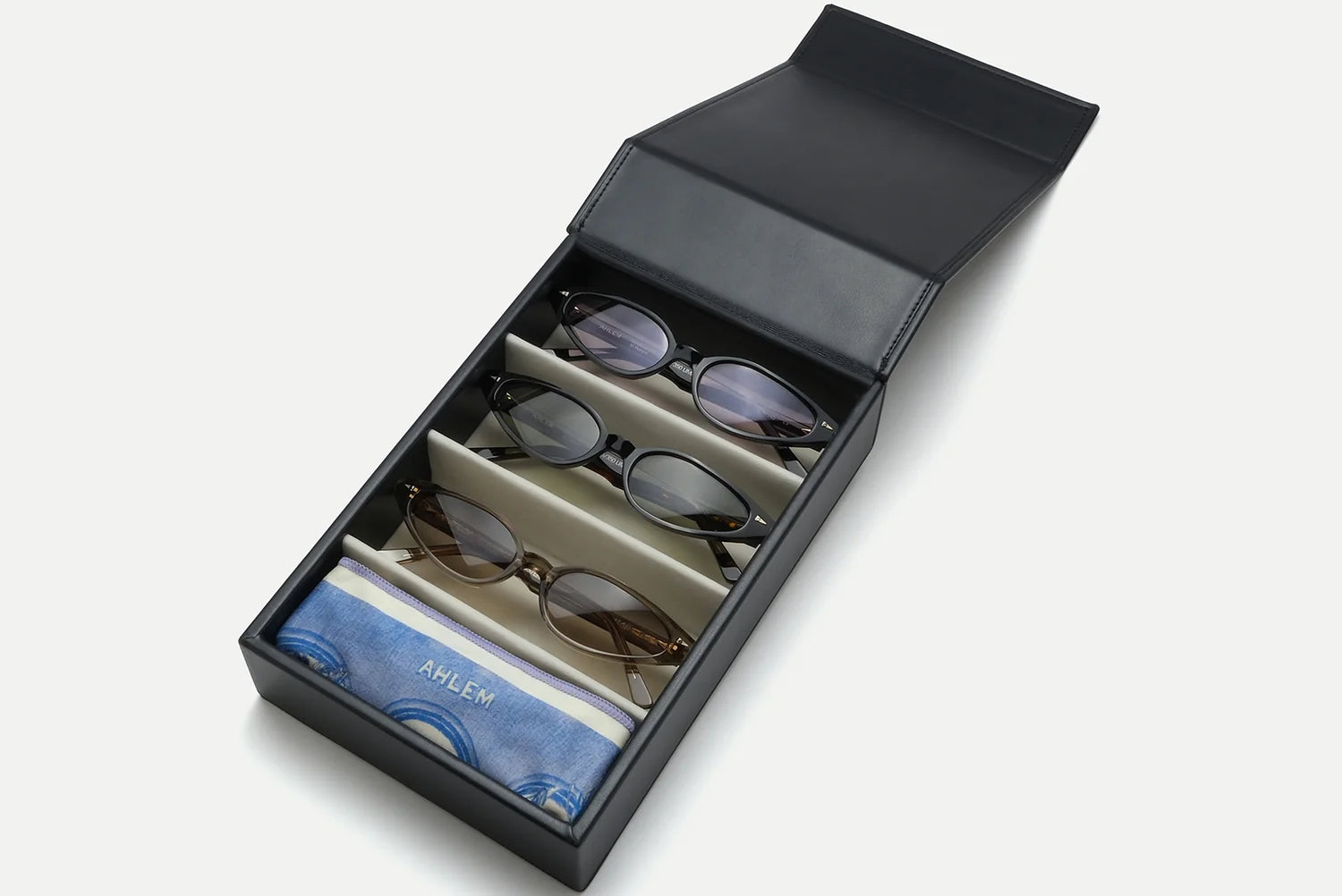 The open AHLEM Voyager Collection: 4-Unit Travel Case by AHLEM displays three pairs of sunglasses, each secured in its own slot, along with a blue microfiber cleaning cloth labeled “AHLEM” positioned at the bottom. All items are neatly organized inside the black travel case.