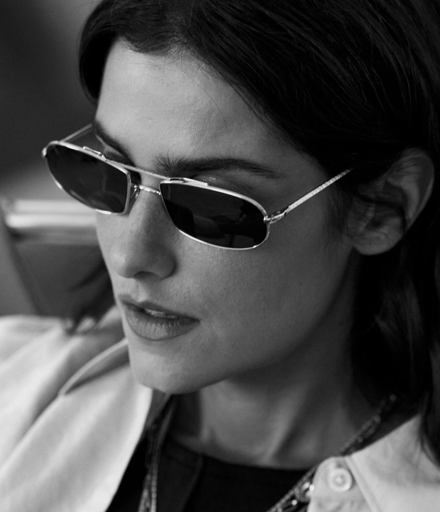 A woman with wavy dark hair wears AHLEM Guerin slim, rectangular unisex sunglasses from AHLEM, handcrafted in France, along with a light-colored jacket. The black-and-white photo captures her looking down with a calm, thoughtful expression.