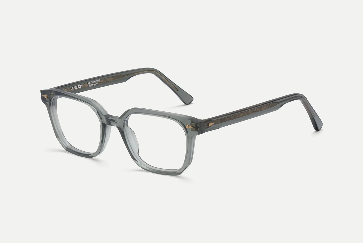 The AHLEM Houdon by AHLEM features clear gray, square-shaped optical glasses with thick frames, expertly handcrafted from slightly transparent acetate. Decorative accents on the arms complement the premium steel hardware, creating a refined balance of elegance and durability. The eyewear is showcased against a plain white background.