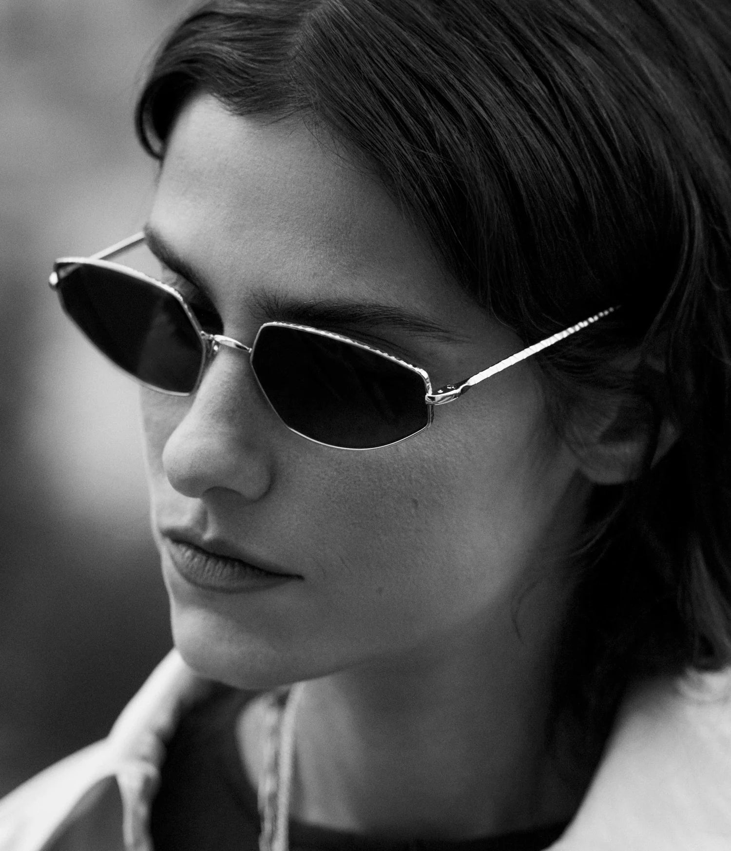 Black and white photo of a person with straight dark hair, wearing AHLEM Yvonne sunglasses handcrafted in France and a light-colored jacket, looking downward with a neutral expression.