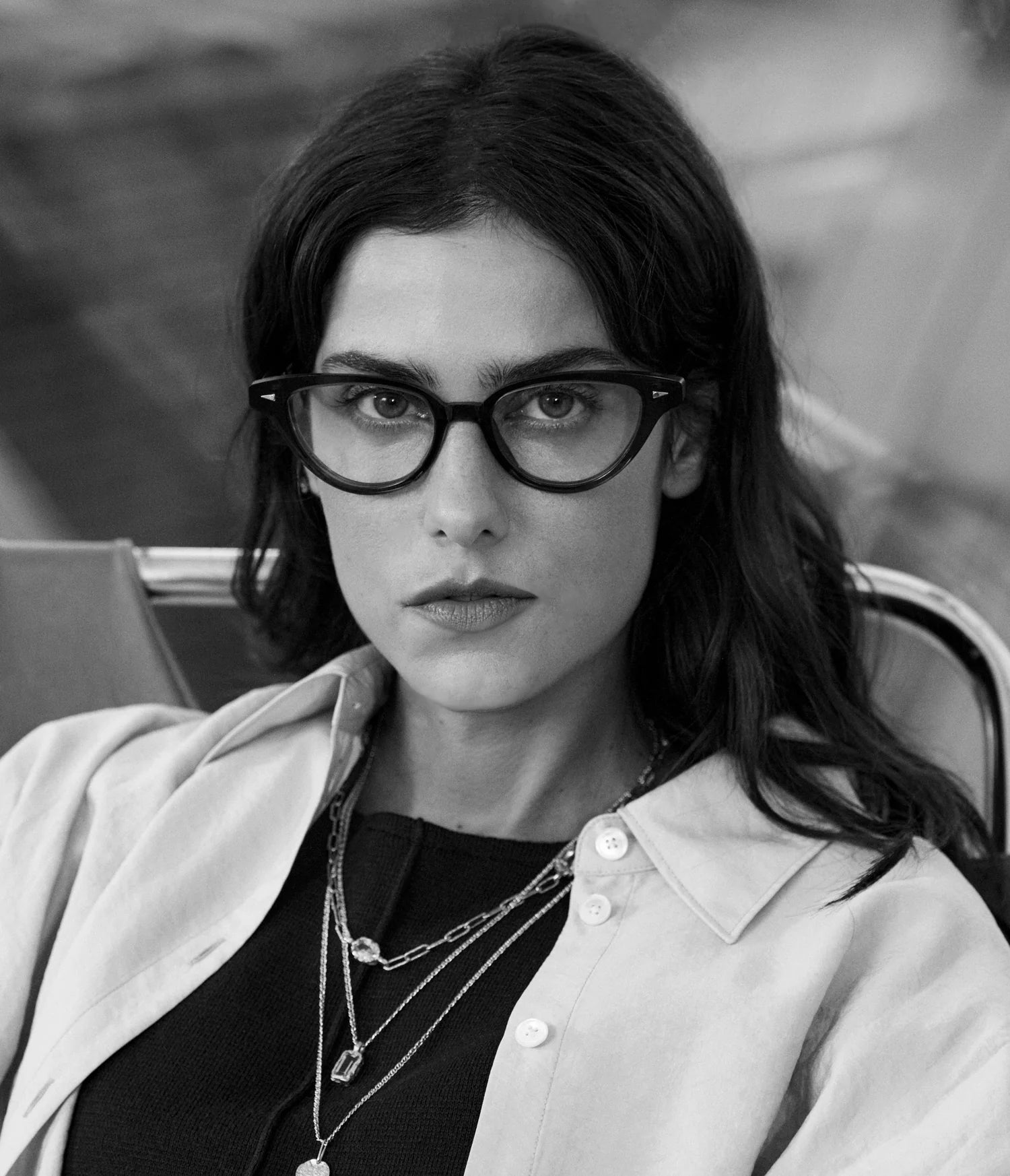 Black and white photo of a woman with long dark hair, wearing handcrafted in France AHLEM Lilas cat-eye optical glasses by AHLEM, layered necklaces, a dark top, and a light jacket, sitting on a chair and looking directly at the camera with a neutral expression.