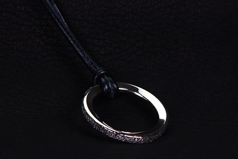 A silver-toned metal ring pendant with a textured, glittery outer edge, hanging from a black cord against a dark textured background.