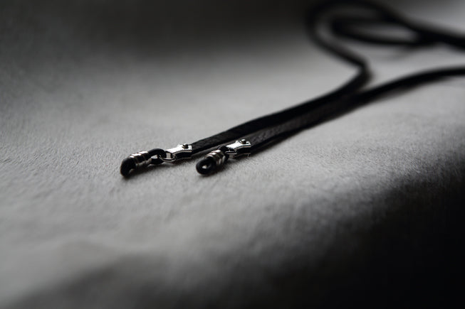 Close-up of two DIFFUSER SILVER RIVETED GLASS CORDS by DIFFUSER, featuring black cords with sterling silver fittings, lying on a soft, light grey surface with a blurred background.