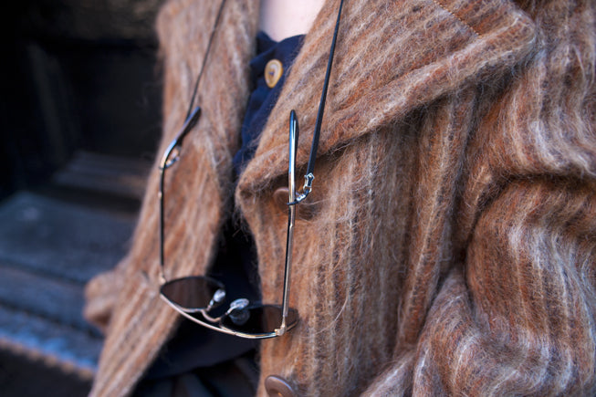 A close-up of a person wearing a fuzzy brown striped coat and a dark shirt, with sunglasses hanging from a DIFFUSER SILVER RIVETED GLASS CORD by DIFFUSER around their neck.