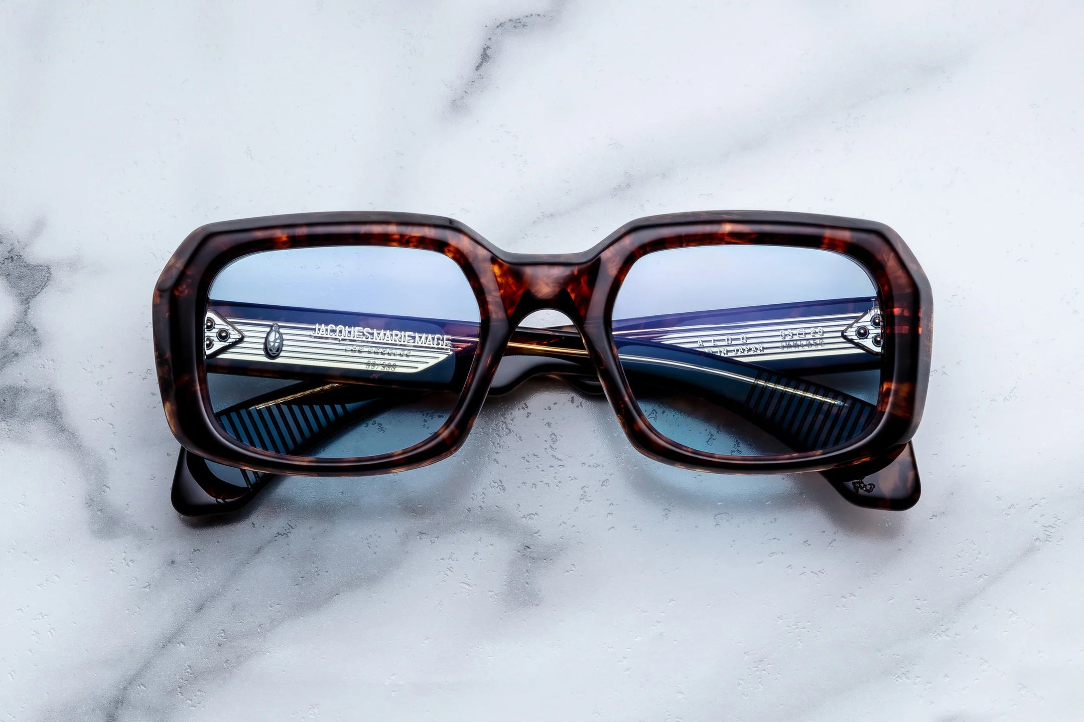 Jacques Marie Mage Aldo eyeglasses by Jacques Marie Mage showcase brown tortoiseshell rectangular frames with a bold, thick silhouette, resting on a white marble surface. Silver accents gleam on the arms, while reflected light highlights the elegant craftsmanship and timeless style of these spectacles.