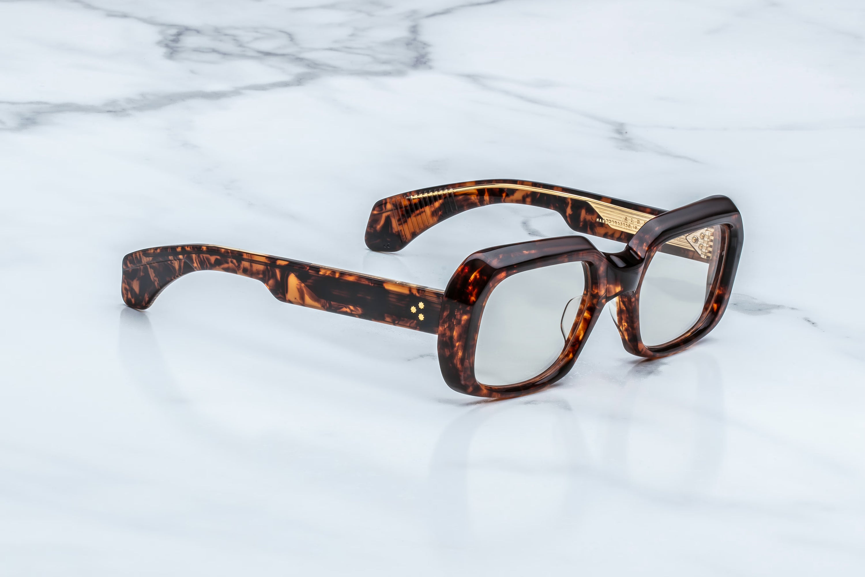 The Jacques Marie Mage Aldo sunglasses by Jacques Marie Mage feature tortoiseshell-patterned, thick square frames and slightly tinted lenses, expertly crafted and elegantly displayed on a white marble surface.