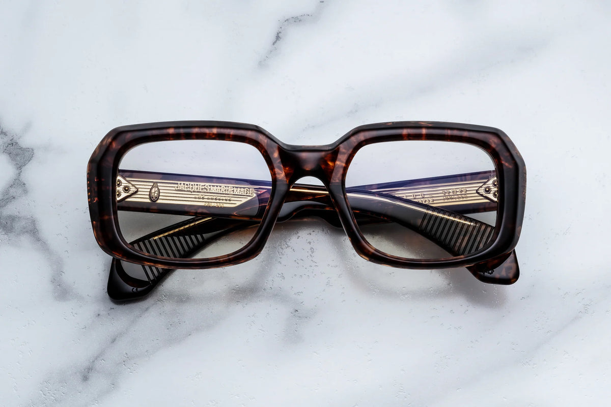 Jacques Marie Mage Aldo rectangular spectacles with thick tortoiseshell frames rest on a white marble surface, showcasing Jacques Marie Mage’s exquisite craftsmanship as the temples cross behind the lenses.
