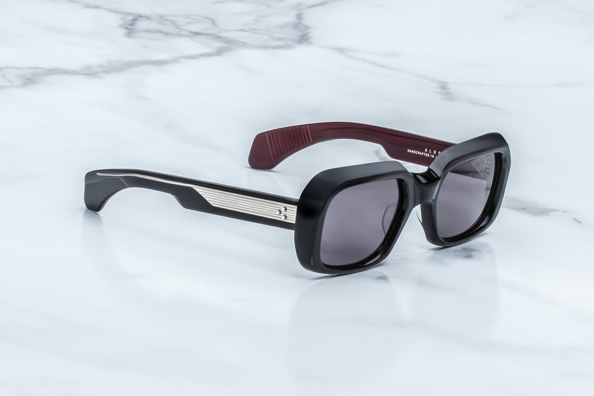 The Jacques Marie Mage Aldo sunglasses by Jacques Marie Mage feature bold black square frames with dark lenses, silver accents on the temples, and a maroon inner side, resting elegantly on a white marble surface—these spectacles showcase timeless style paired with exceptional craftsmanship.