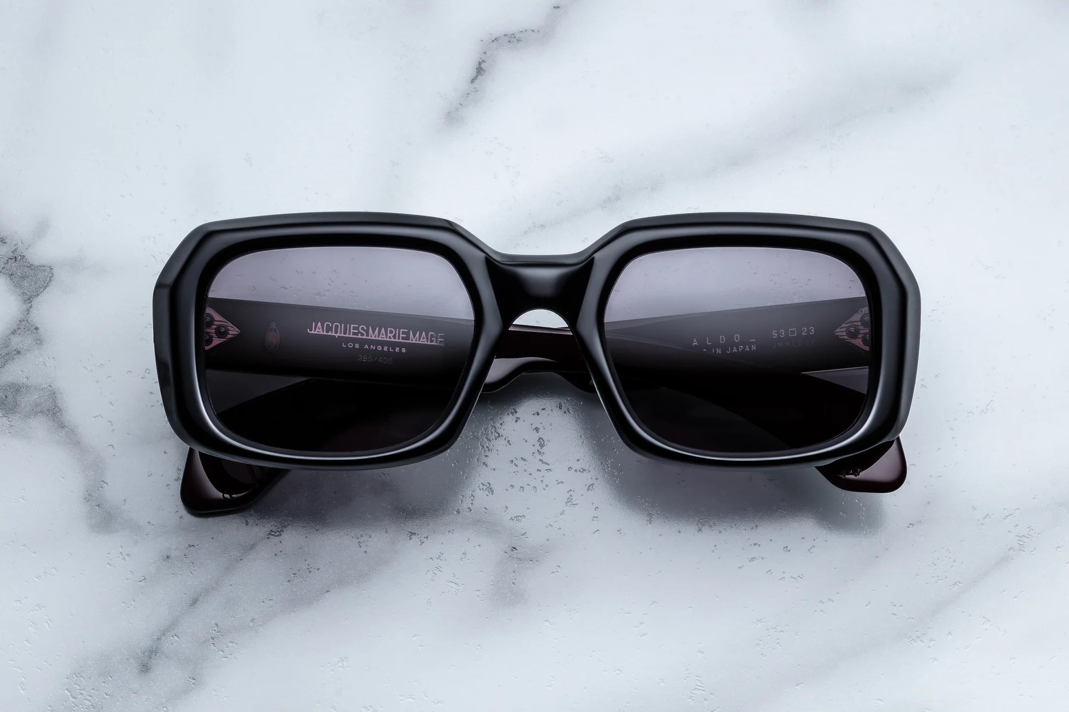 The Jacques Marie Mage Aldo sunglasses, featuring black square frames and dark lenses, rest on a white marble surface that reflects the light. Exemplifying superior craftsmanship, these spectacles display the “Jacques Marie Mage” brand name on the inside of the left lens, embodying a legacy of timeless fashion.
