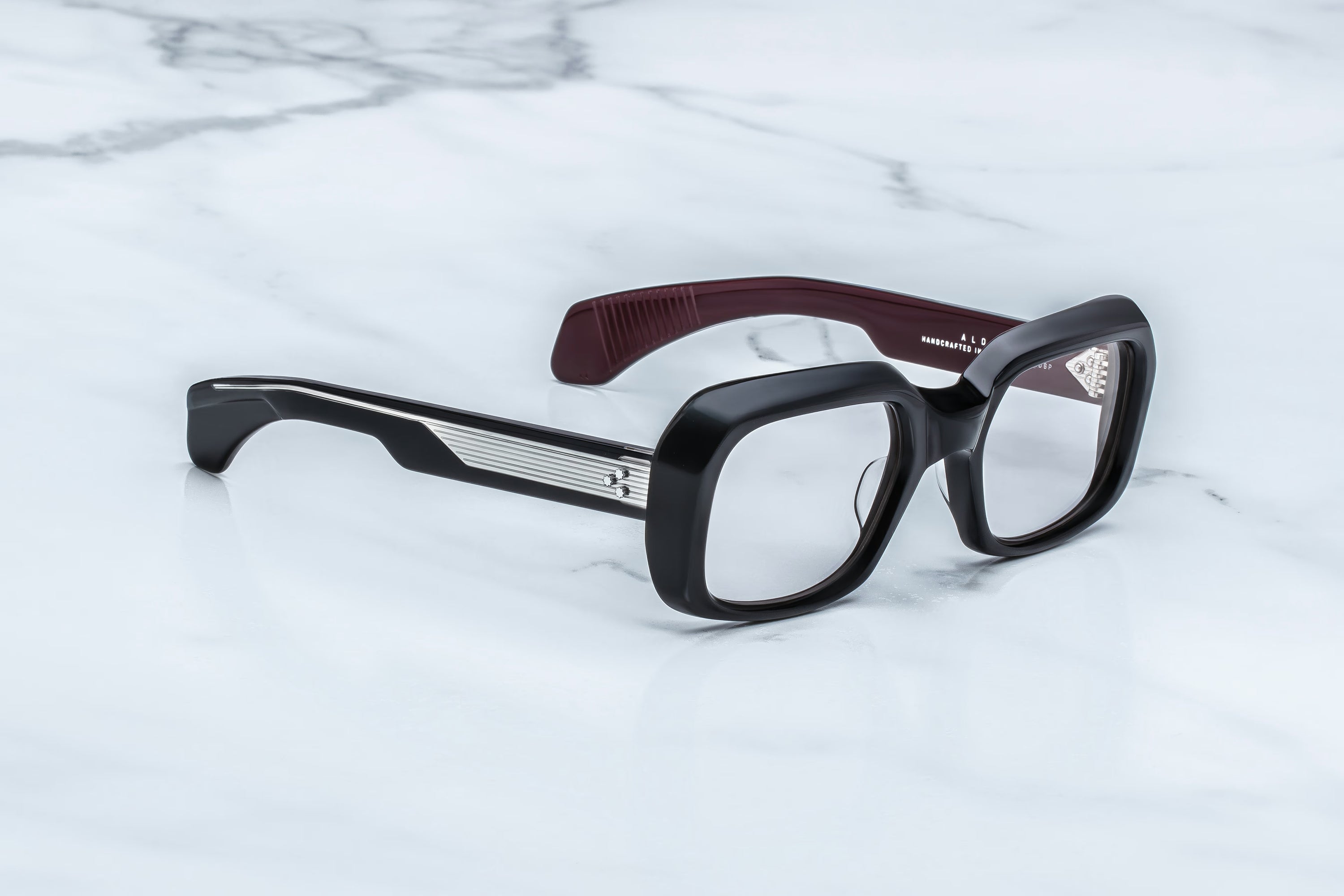 Jacques Marie Mage Aldo spectacles by Jacques Marie Mage, featuring bold black square frames with thick rims and sophisticated dark maroon arms, rest elegantly on a white marble surface—a true testament to timeless fashion and expert craftsmanship.