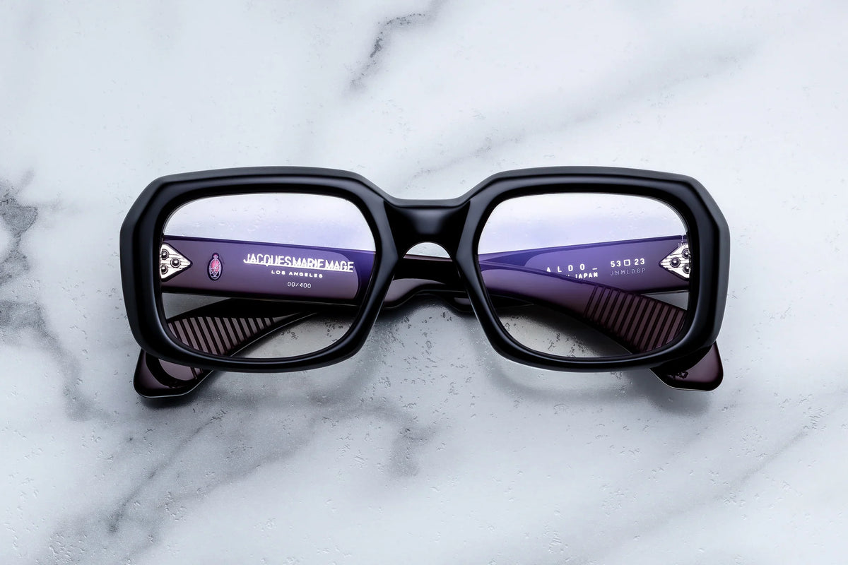 Jacques Marie Mage Aldo eyeglasses featuring bold black square frames rest atop a white marble surface, exemplifying the refined craftsmanship and timeless elegance for which Jacques Marie Mage is renowned. Reflections in the lenses reveal distinctive brand details and text on the inner arms of these classic spectacles.