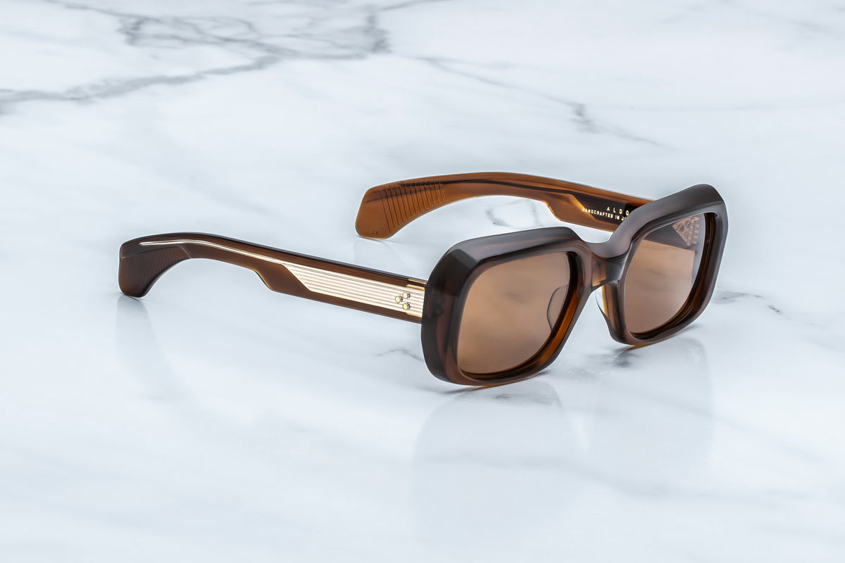 The Jacques Marie Mage Aldo sunglasses by Jacques Marie Mage, featuring brown square lenses, thick frames, and gold accents on the temples that highlight exceptional craftsmanship, rest elegantly on a white marble surface.