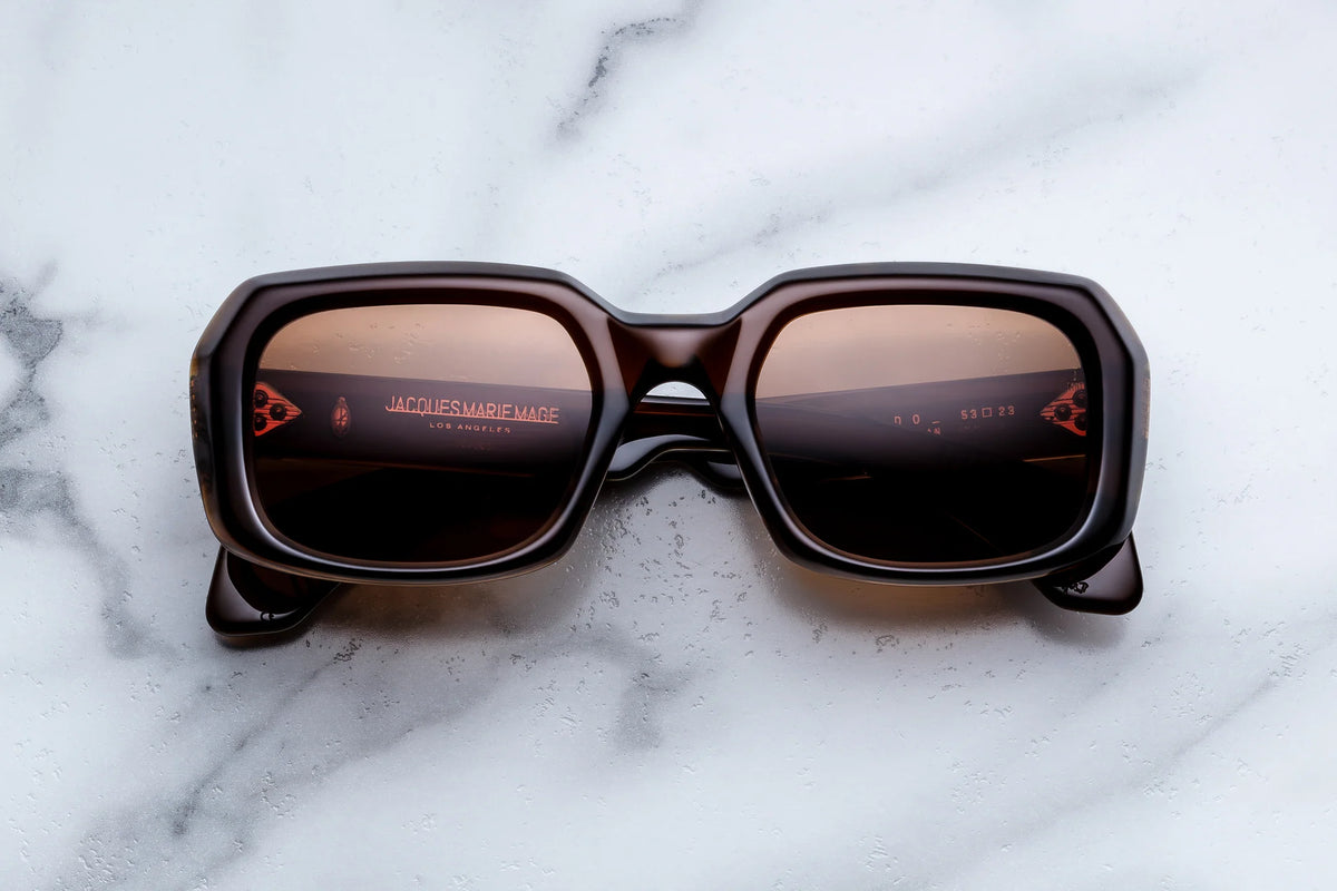 Jacques Marie Mage Aldo brown rectangular sunglasses with dark lenses rest on a white marble surface. Showcasing exceptional craftsmanship, the name Jacques Marie Mage is faintly visible on the inside of the left lens.