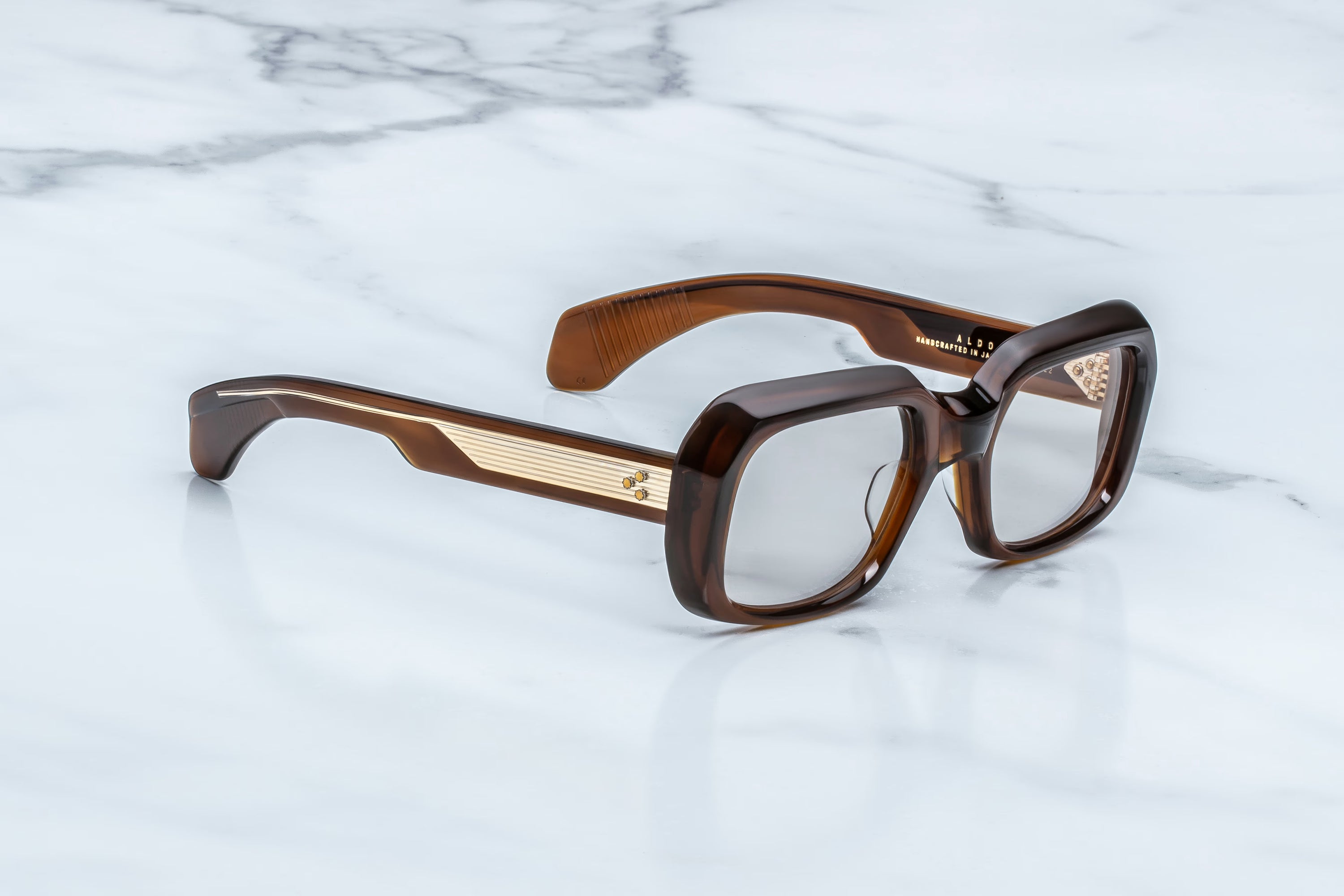 The Jacques Marie Mage Aldo by Jacques Marie Mage: chunky, square-framed brown spectacles with gold-striped detailing on the arms, resting atop a white marble surface—an elegant nod to timeless fashion and exquisite craftsmanship.