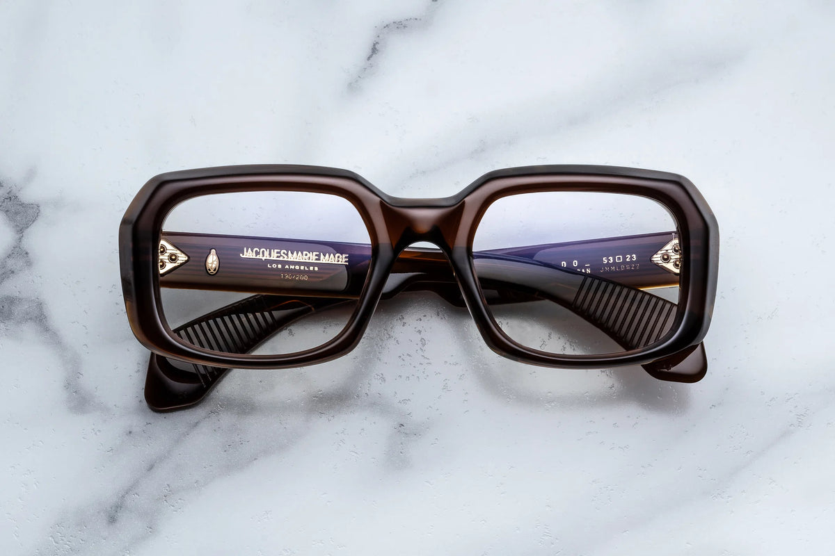 Jacques Marie Mage Aldo square spectacles with thick brown frames are positioned lens-up on a white marble surface, highlighting the Jacques Marie Mage branding on the inside of the right temple—a testament to exceptional craftsmanship and timeless style.