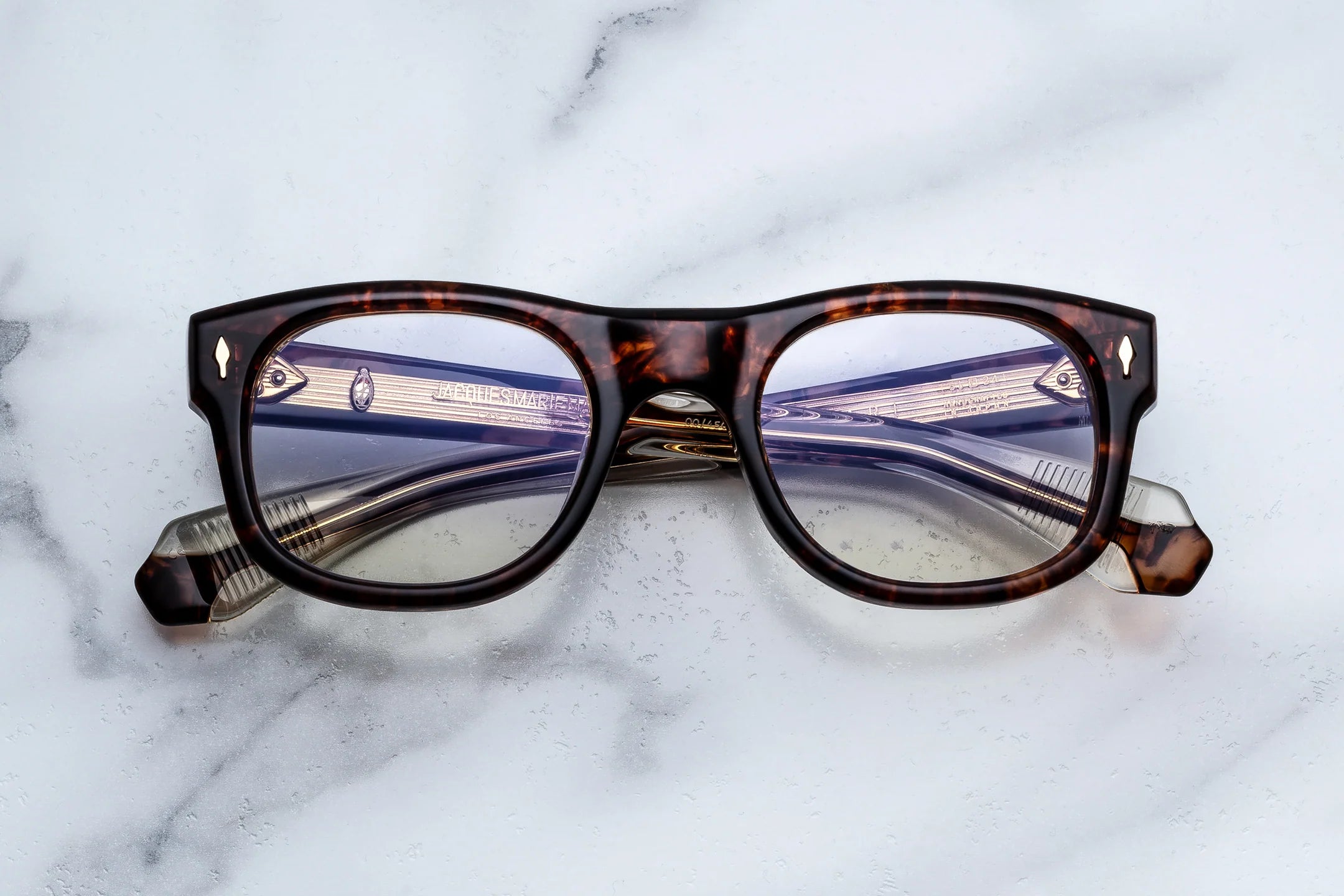 A pair of classic-style eyeglasses with a dark brown and amber tortoiseshell acetate frame, resting on a white marble surface.