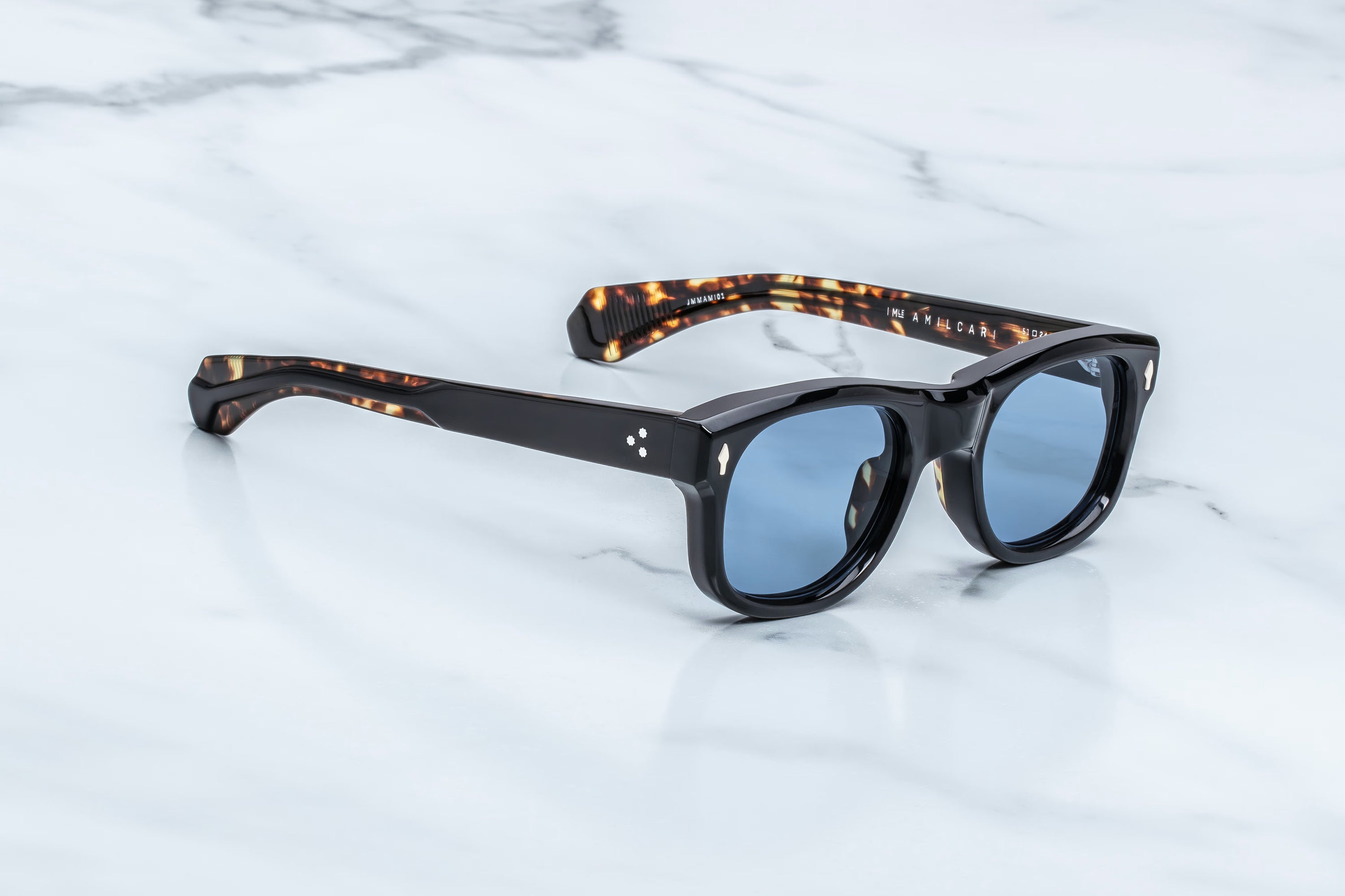 A pair of sunglasses with black acetate frames, blue-tinted lenses, and tortoiseshell-patterned temples, resting on a white marble surface.