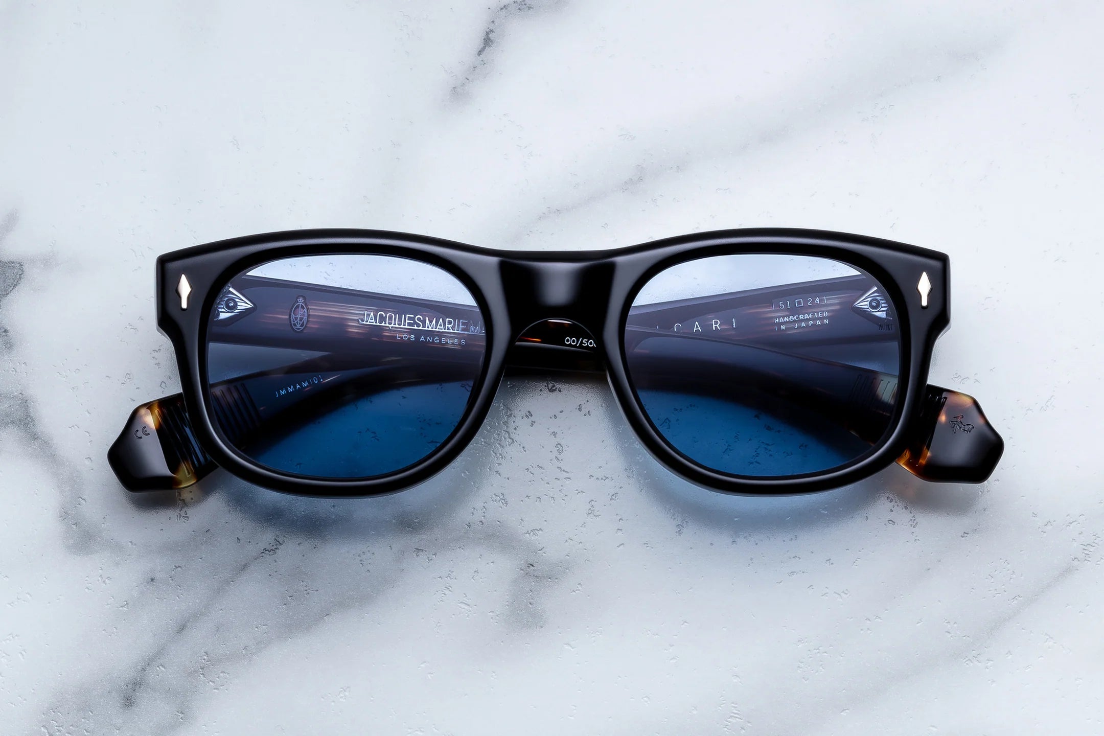 A pair of black square sunglasses with blue-tinted lenses and tortoiseshell temple tips, photographed from above on a white marble surface.