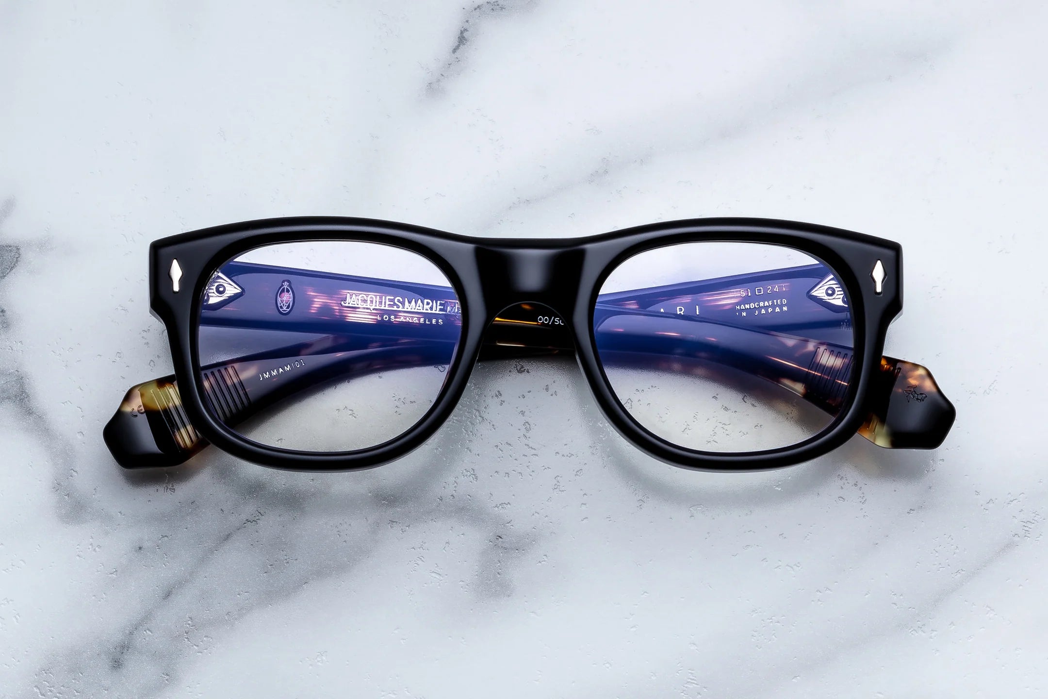 A pair of black square eyeglasses with tortoiseshell temple tips, displayed flat on a white marble surface. The lenses have a blue tint and reflect the photographer's surroundings.