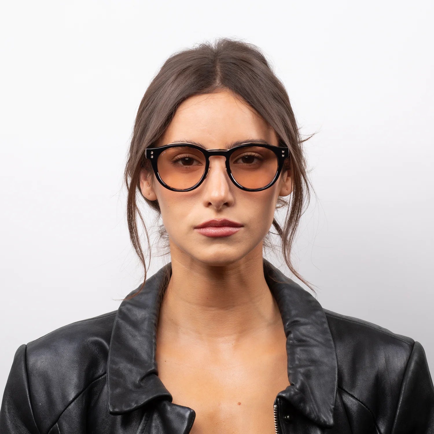 A woman with brown hair wearing black round eyeglasses with amber-tinted lenses and a black leather jacket, photographed against a light gray background.