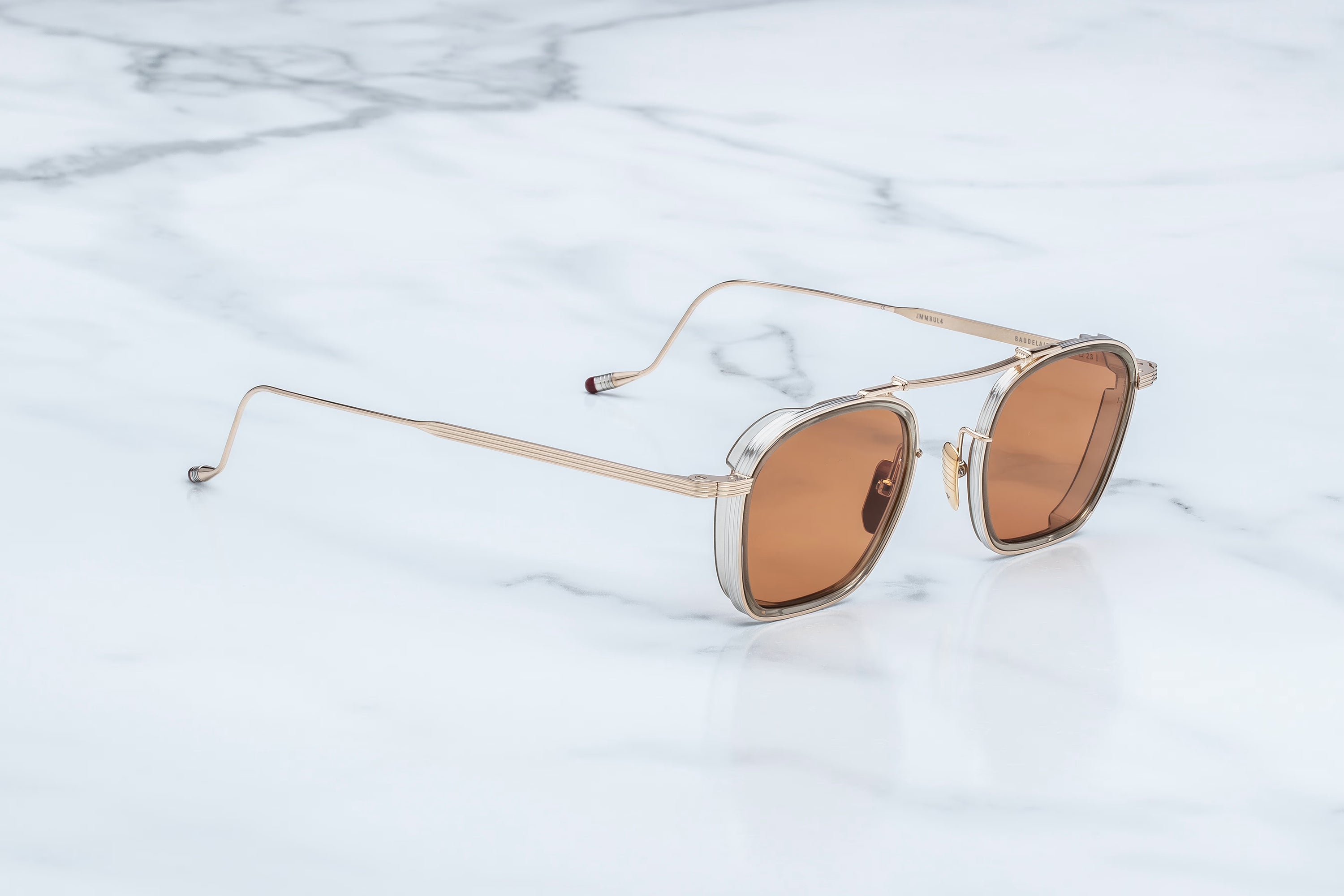 Jacques Marie Mage Baudelaire sunglasses by Jacques Marie Mage, featuring gold frames and brown lenses, rest on a white marble surface—highlighting the allure of handcrafted eyewear.