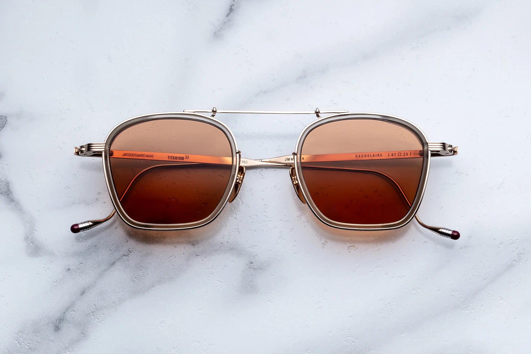 Jacques Marie Mage Baudelaire sunglasses by Jacques Marie Mage, featuring square-shaped gold frames and brown tinted lenses, are displayed on a white marble surface, highlighting the brand's handcrafted eyewear that embodies elegance and style.