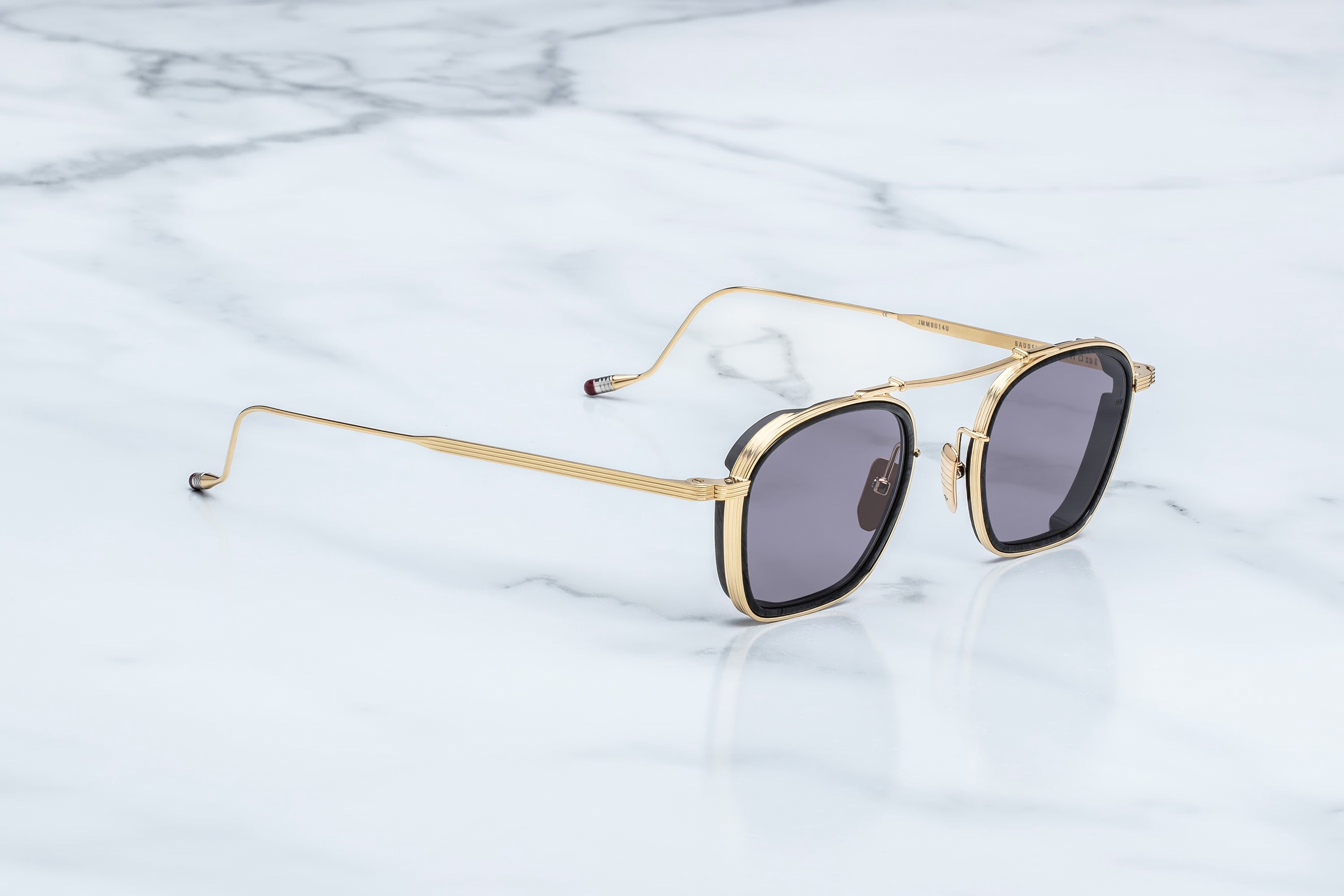 The Jacques Marie Mage Baudelaire sunglasses by Jacques Marie Mage, featuring gold frames and dark lenses, are displayed on a white marble surface. Angled slightly to the left with their slender, elegant arms extended and intricate detailing along the frame edges, these unique spectacles perfectly embody timeless style.