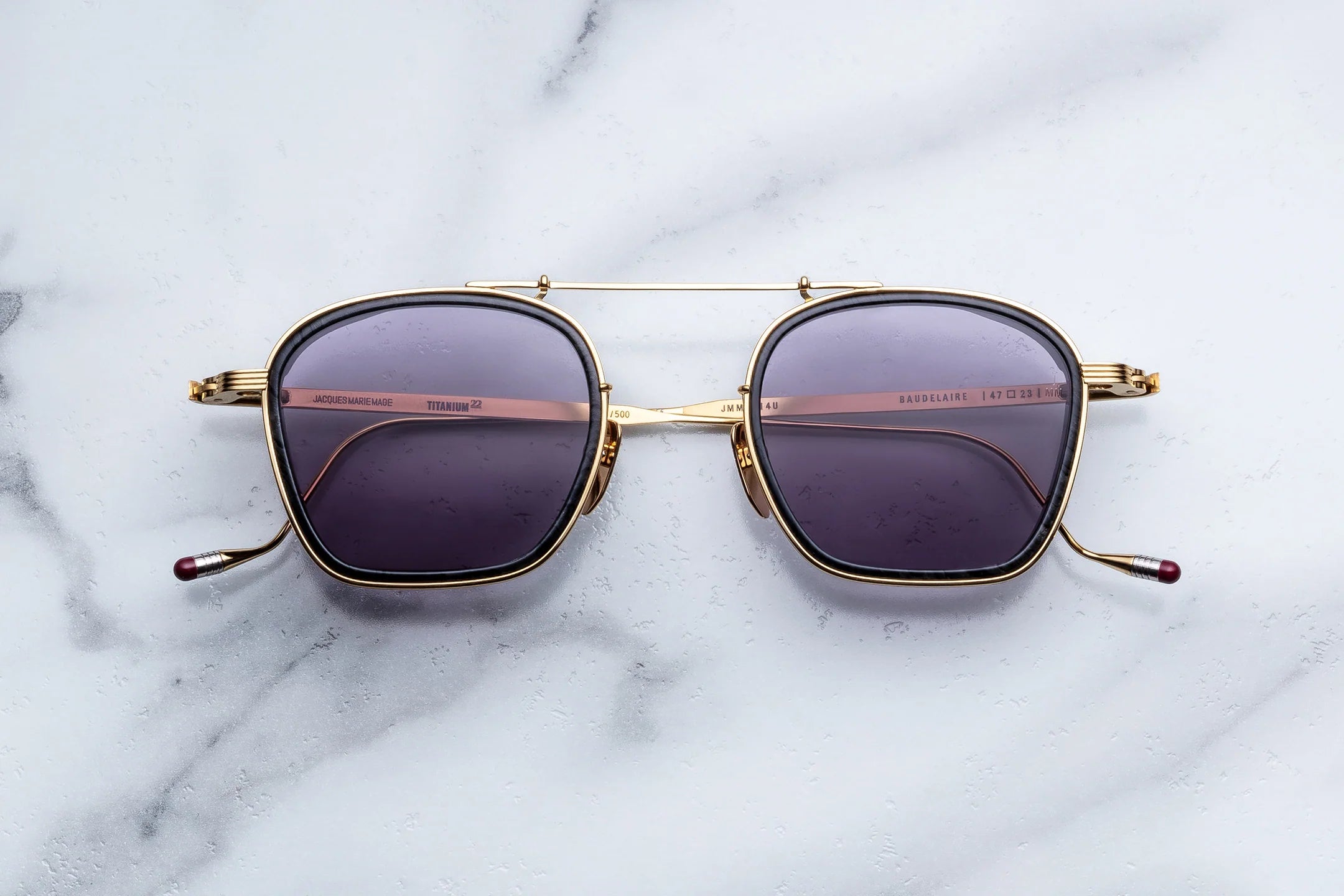 A pair of Jacques Marie Mage Baudelaire sunglasses, featuring square gold frames and purple tinted lenses, rests on a white marble surface—an elegant example of handcrafted eyewear by Jacques Marie Mage.