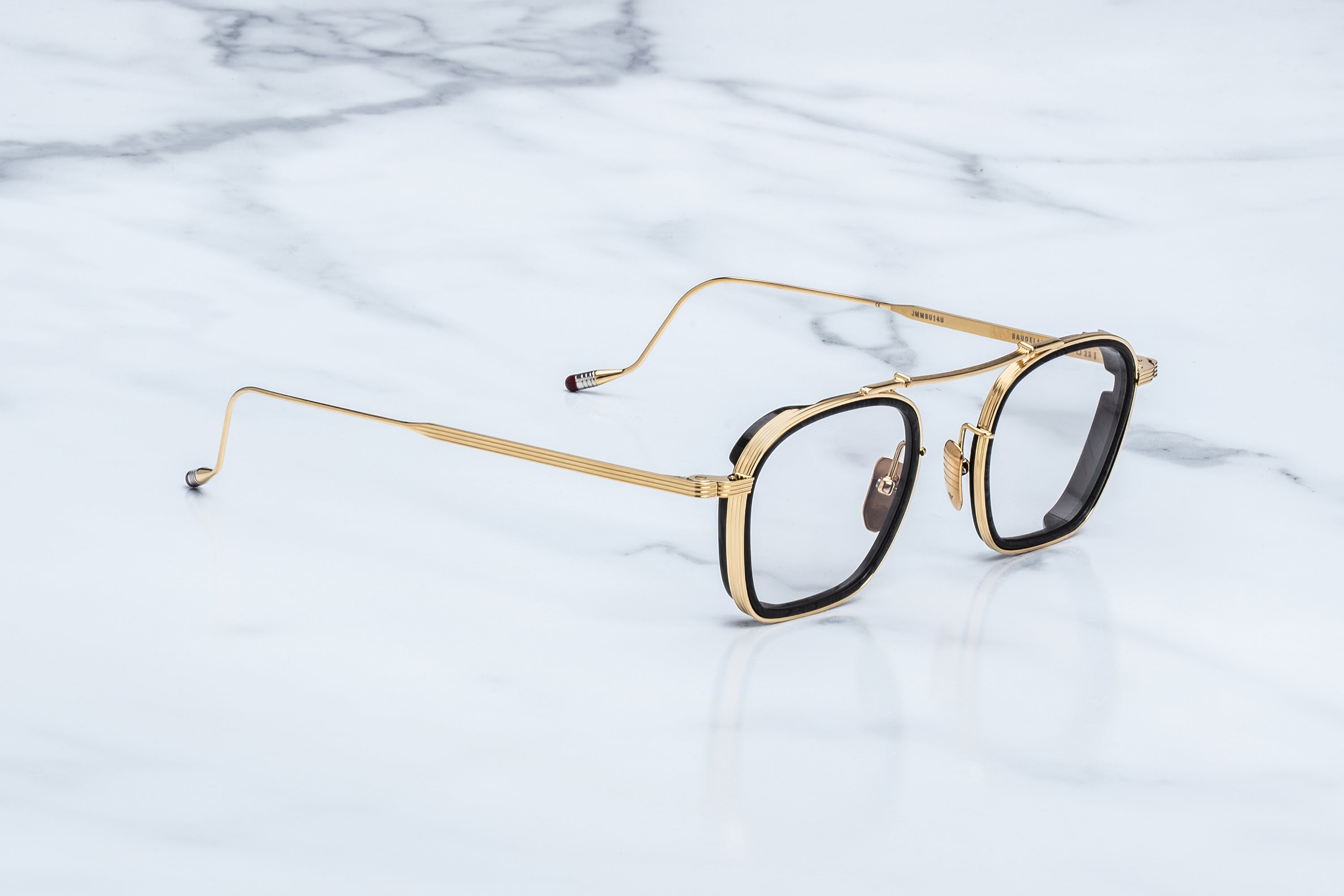 Jacques Marie Mage Baudelaire eyeglasses by Jacques Marie Mage, featuring gold-rimmed square frames with black accents and thin gold temples, rest on a white marble surface. Their rounded edges showcase a timeless design that effortlessly blends elegance and sophistication.
