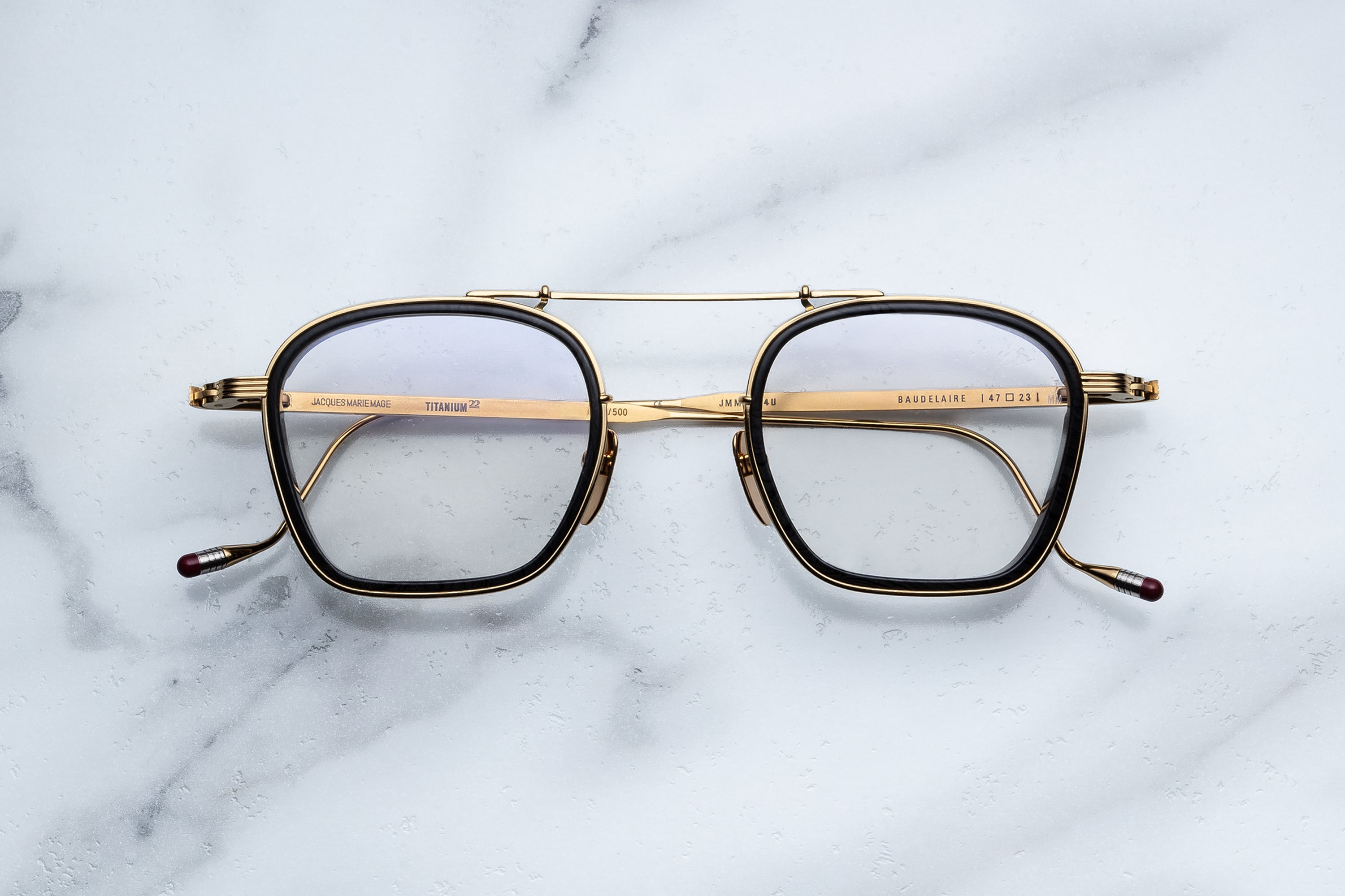 The Jacques Marie Mage Baudelaire eyeglasses by Jacques Marie Mage, featuring a gold and black rectangular double bridge frame with sculptural flourishes, are displayed from above on a white marble surface.