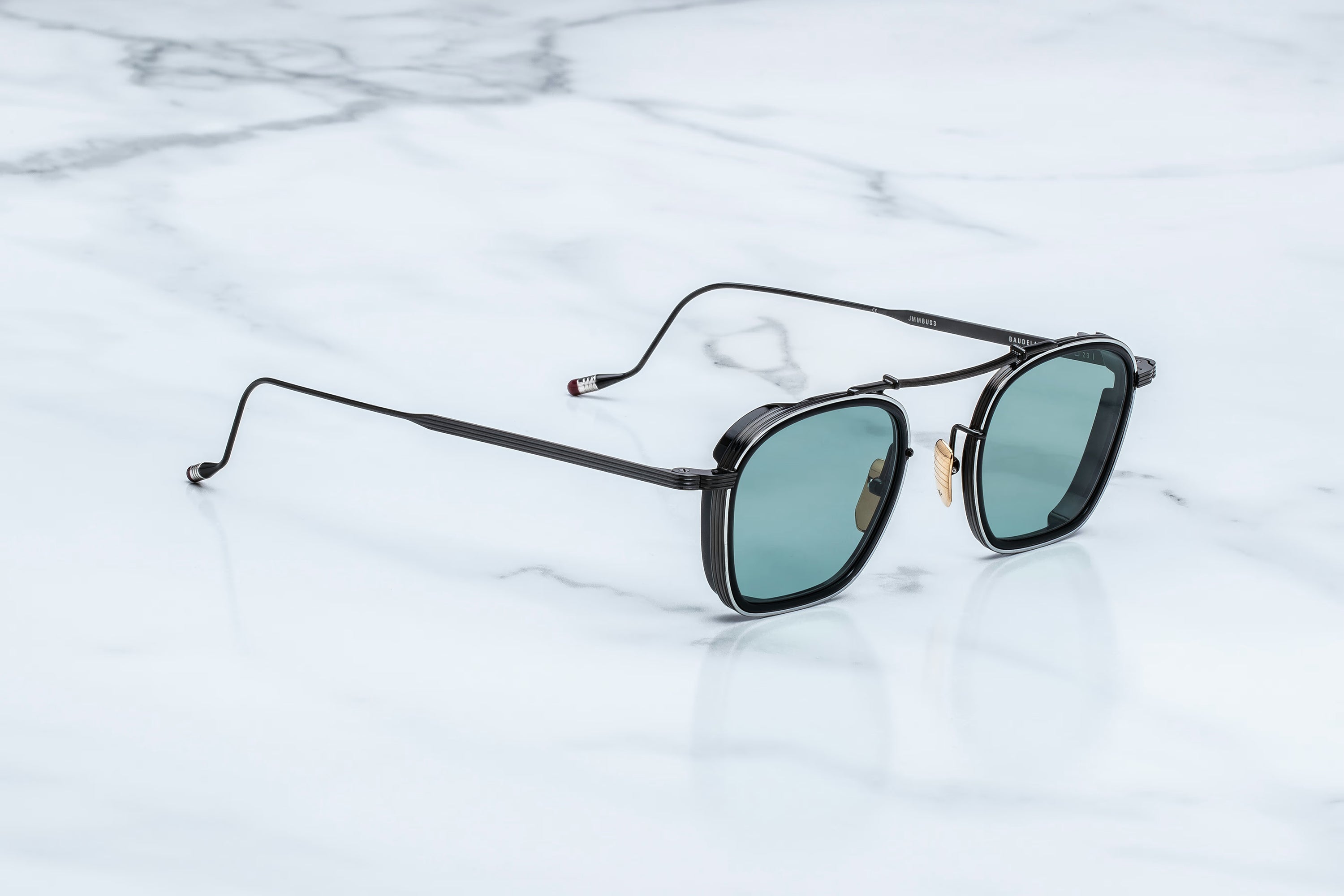 A pair of Jacques Marie Mage Baudelaire sunglasses, featuring classic black frames and green-tinted lenses, rests on a white marble surface.