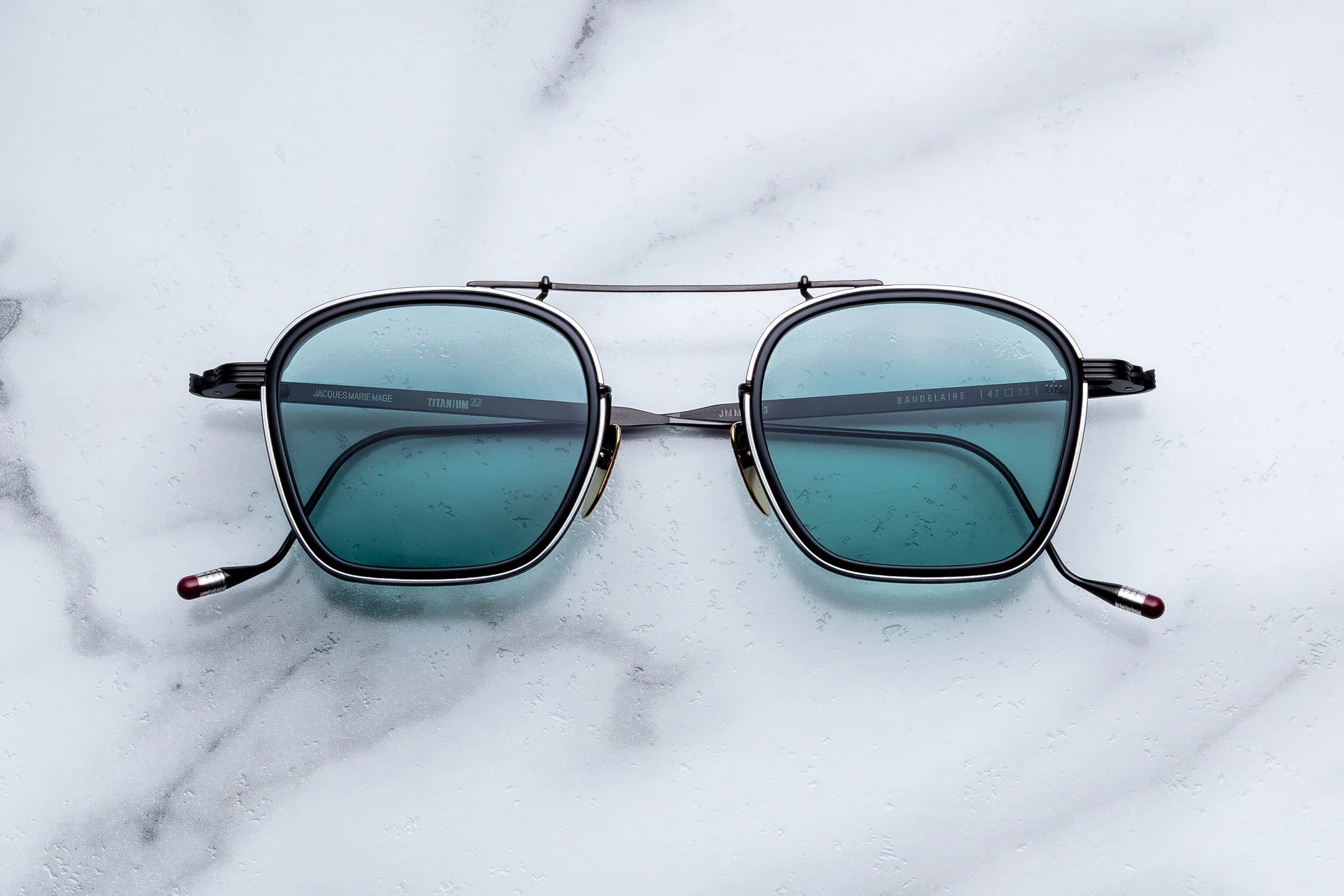 Jacques Marie Mage Baudelaire sunglasses by Jacques Marie Mage, featuring square, teal-tinted lenses and thin black frames with a double bridge, are elegantly displayed on a white marble surface with grey veining, highlighting the distinctive allure of these unique spectacles.