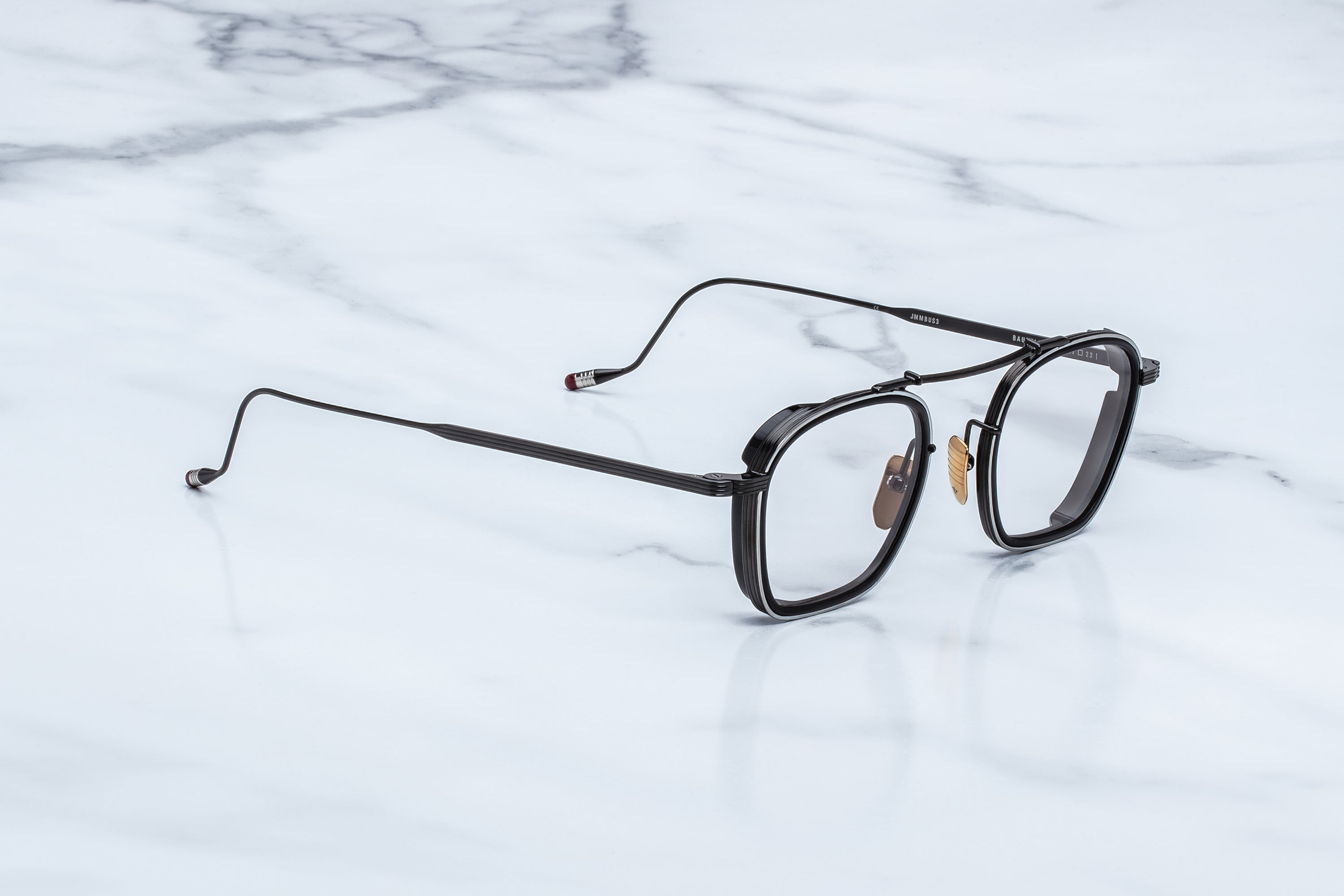 The Jacques Marie Mage Baudelaire eyeglasses by Jacques Marie Mage rest on a white marble surface, showcasing their black round thin metal frames, elegantly curved temples, clear lenses, gold nose pads, and a distinctive handcrafted design that exudes understated elegance.