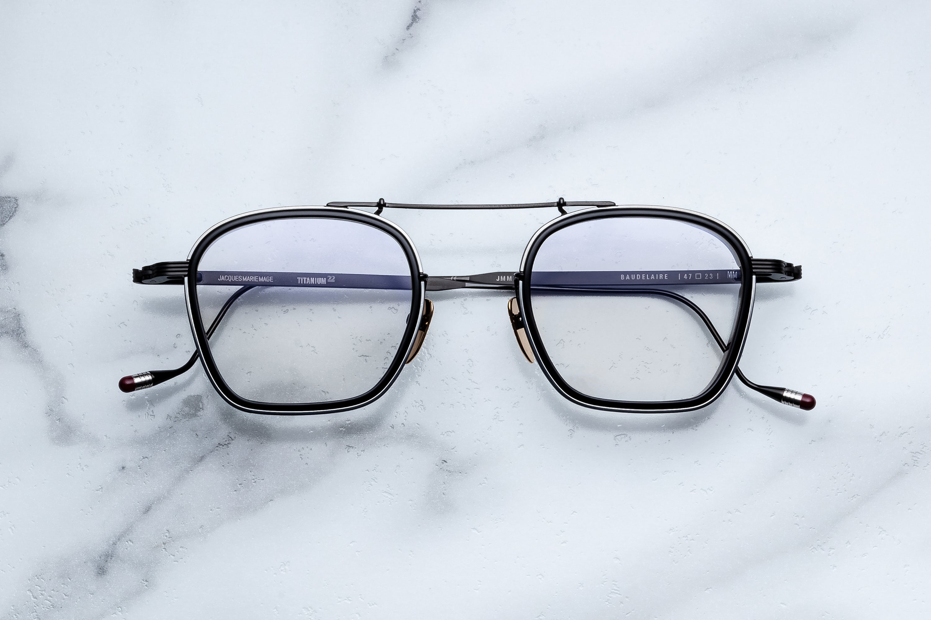 A pair of Jacques Marie Mage Baudelaire eyeglasses by Jacques Marie Mage, featuring black rims and rectangular lenses with a distinctive design, is displayed on a white marble surface.