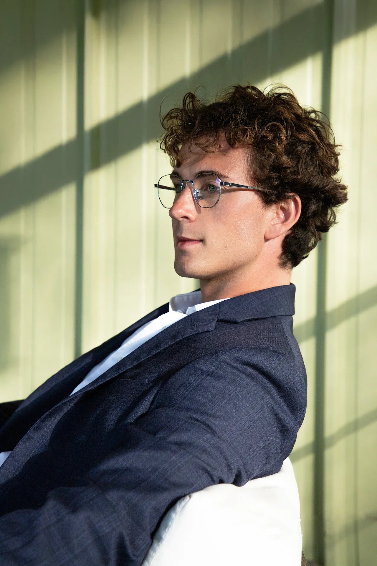 A man with curly brown hair and glasses featuring Leisure Society Bouzy titanium eyeglass frames, wearing a navy suit and white shirt, sits indoors in sunlight, looking to the side. A green paneled wall and shadows are visible in the background.